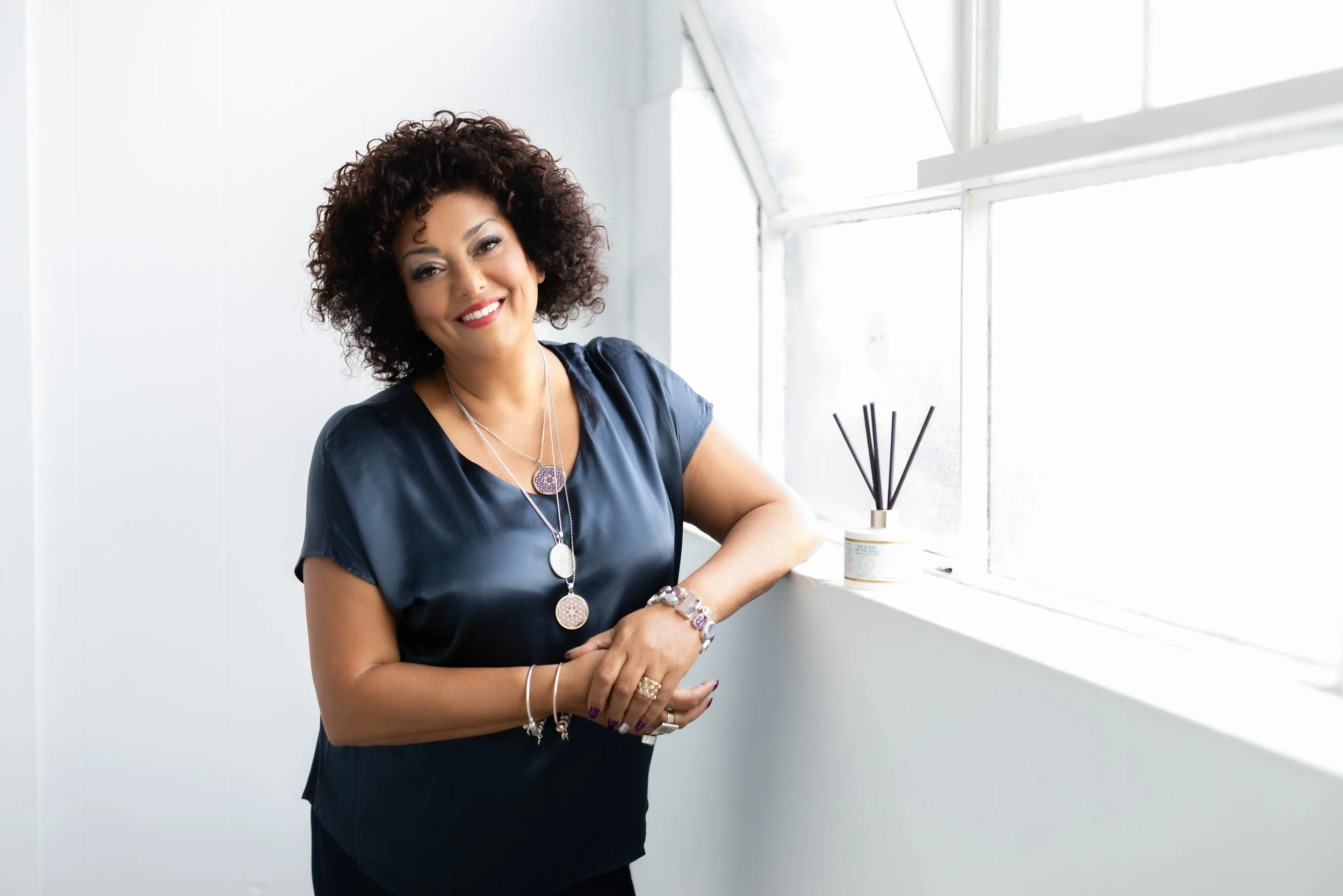 Woman with curly hair smiling and leaning on a windowsill with a reed diffuser on it.