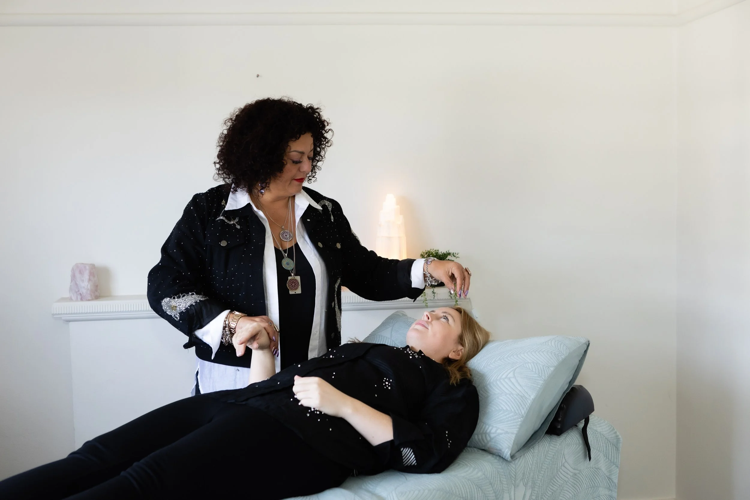 A woman lying on a bed with a black outfit and jewelry, receiving a facial treatment from another woman with curly hair, inside a bright room with minimal decor and a salt lamp.