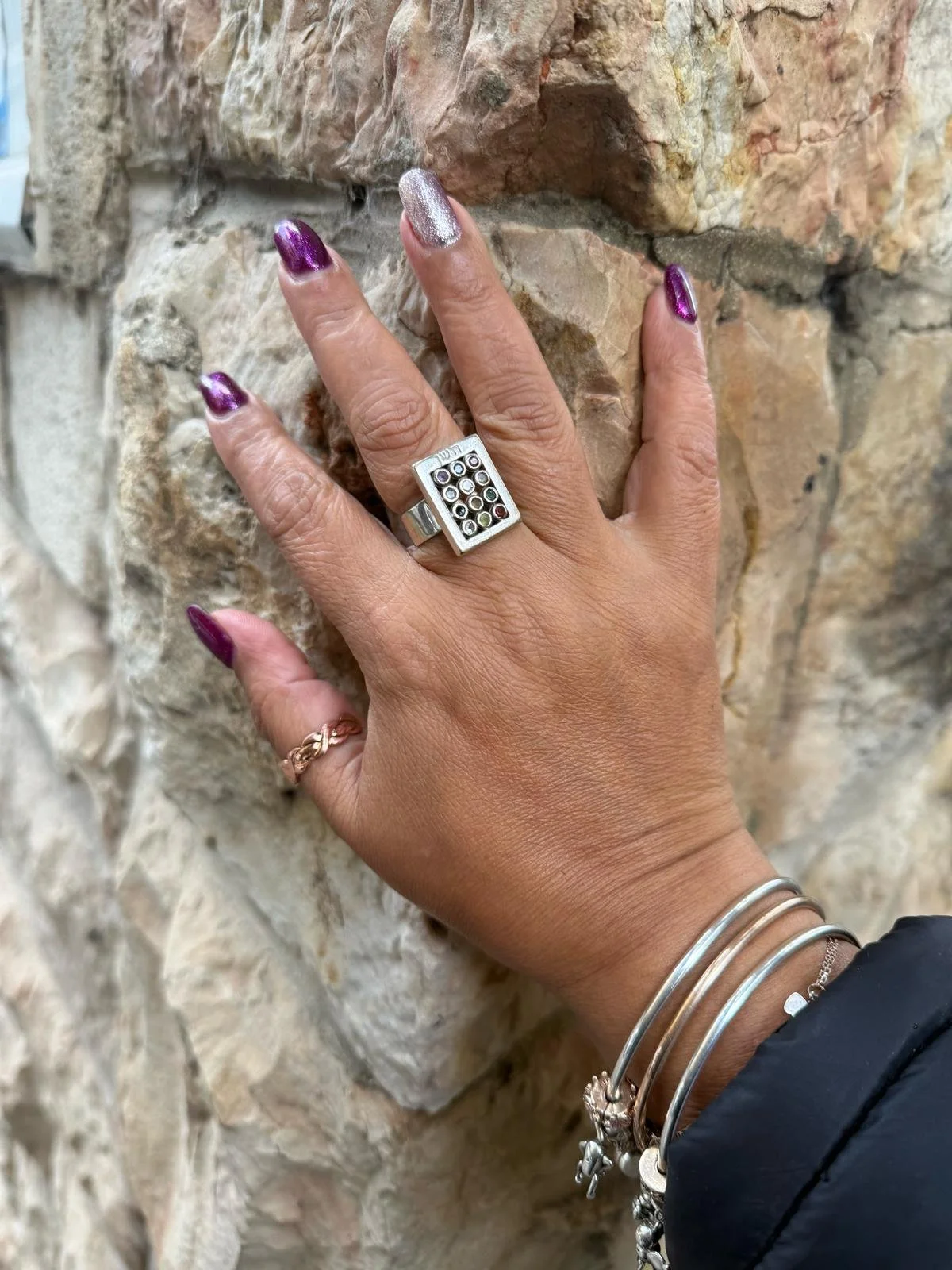 A person's hand with colorful nail polish resting on a stone wall, wearing rings and bracelets.
