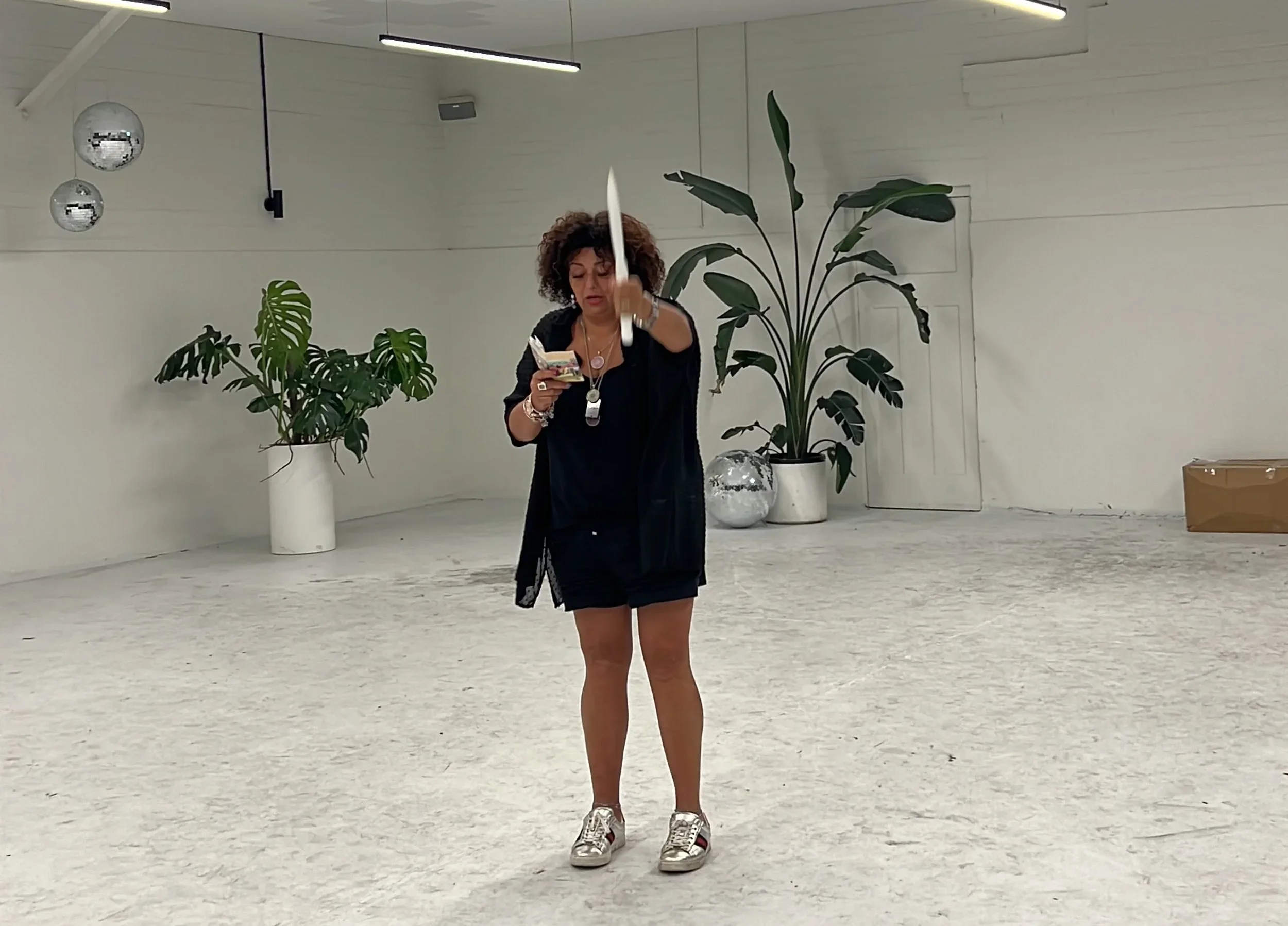 A woman with curly hair wearing a black outfit and sneakers is striking a pose with a baseball bat in a minimalistic white room with large plants and hanging disco balls.