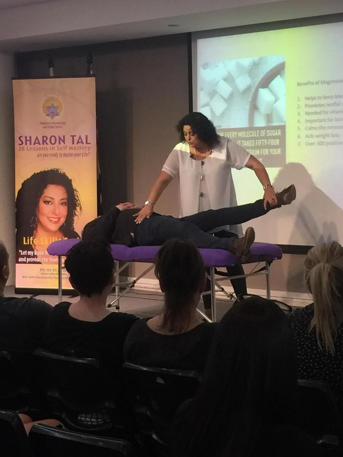 A woman giving a presentation on magnesium benefits, with a person lying on a massage table in front of her. A large poster of the woman with her name, Sharon Tal, is visible to the side.