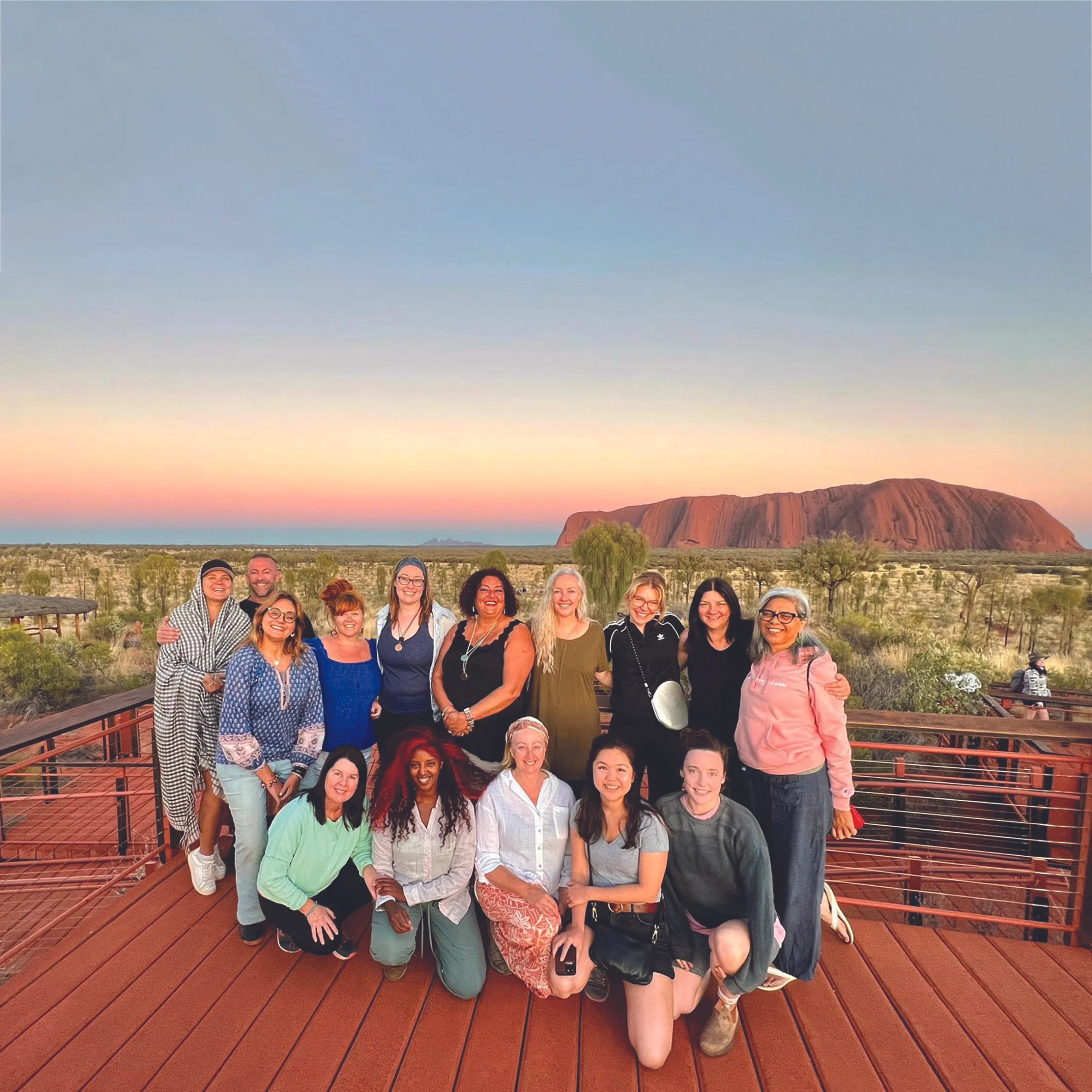 A group of people, mostly women, posing on a wooden observation deck with Uluru rock in the background at sunset.