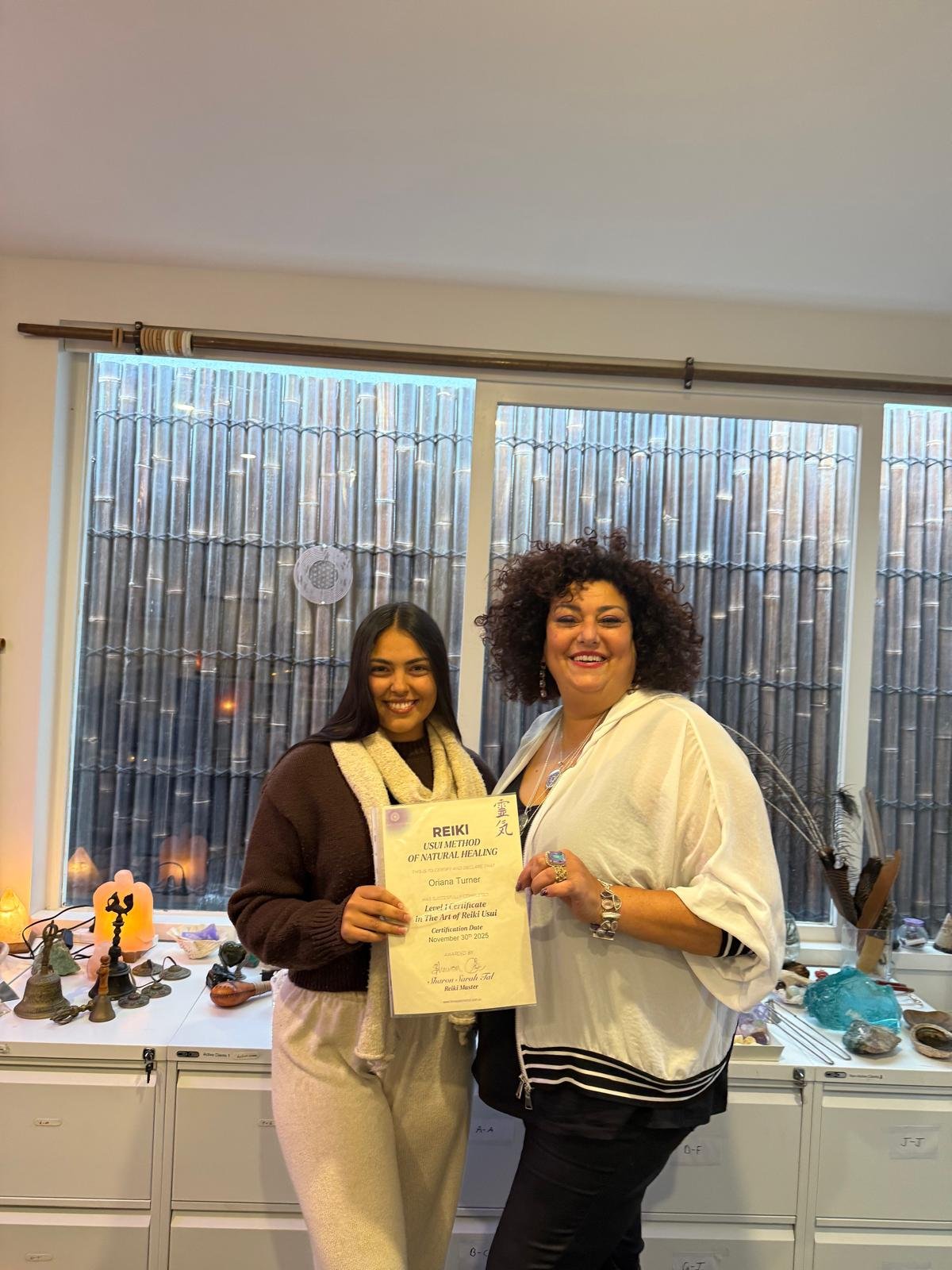 Two women, one holding a Reiki certification, smiling in a room with a table of crystals, statues, and spiritual items, and a large window with bamboo outside.