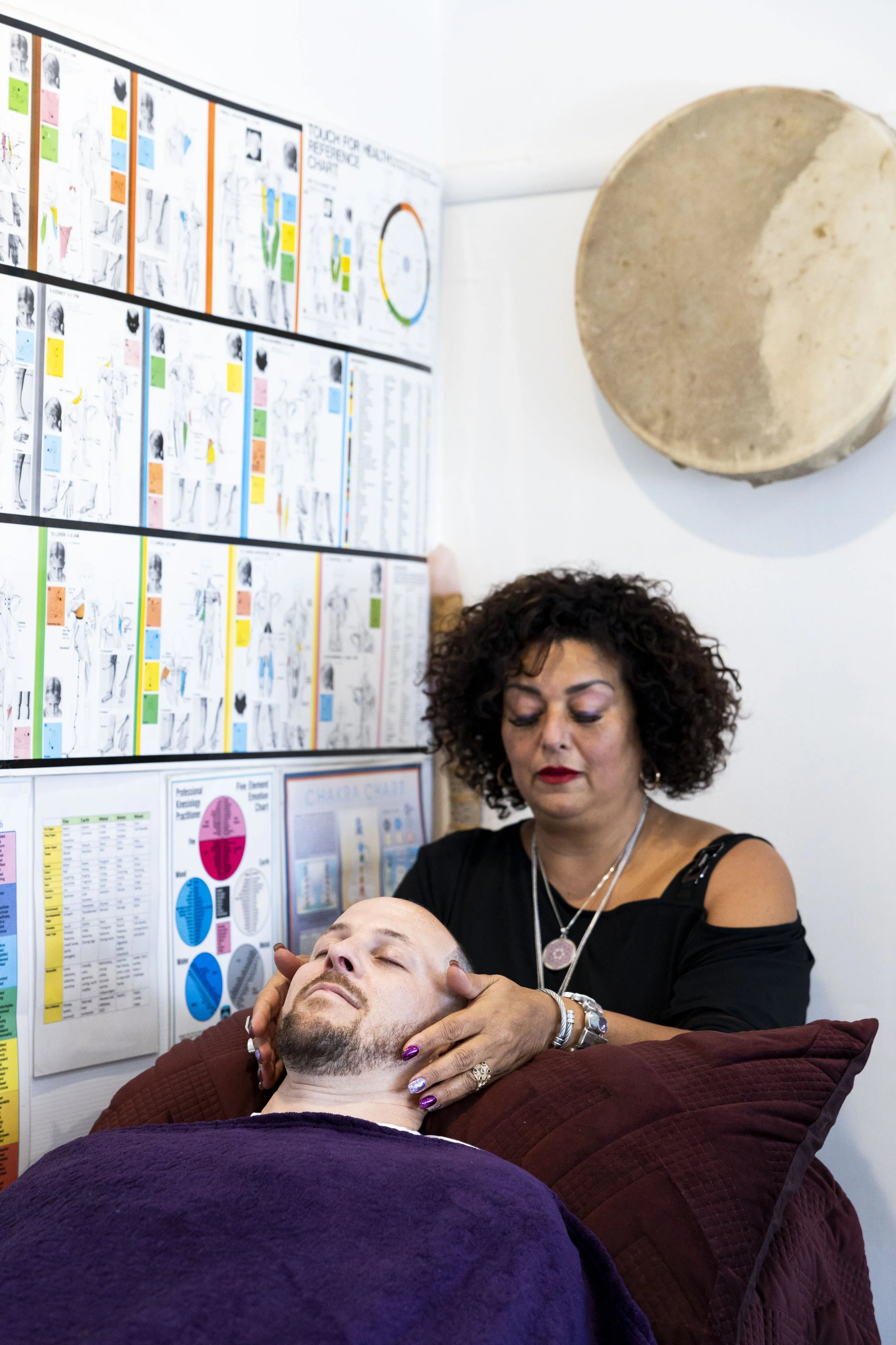 A woman giving a neck massage to a man lying on a cushioned table in an office or therapy room. There are charts and diagrams on the wall behind them.