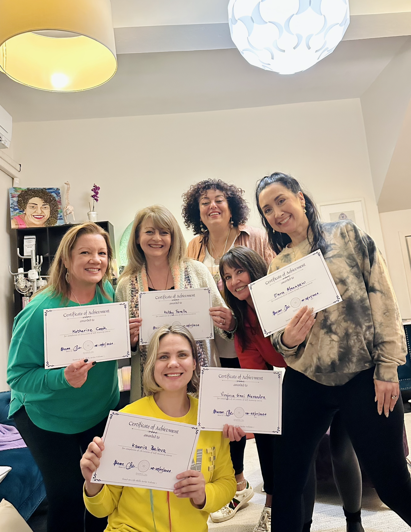 Group of seven women indoors, holding certificates of achievement, smiling, standing in front of a white wall with artwork, plants, and ceiling lights.