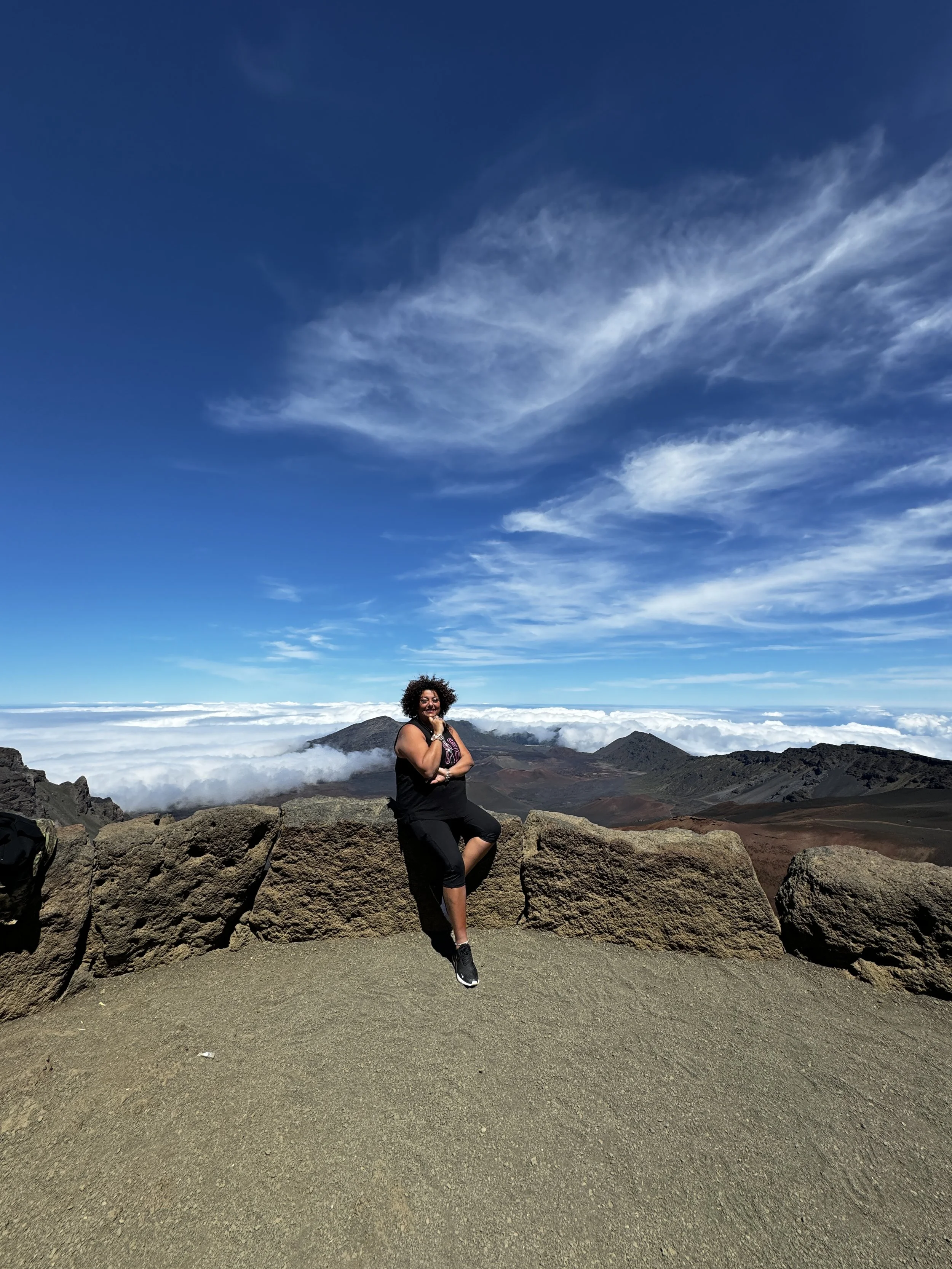 A woman sitting on rocks at a mountain summit with a scenic view of clouds and mountain peaks under a blue sky with wispy clouds.