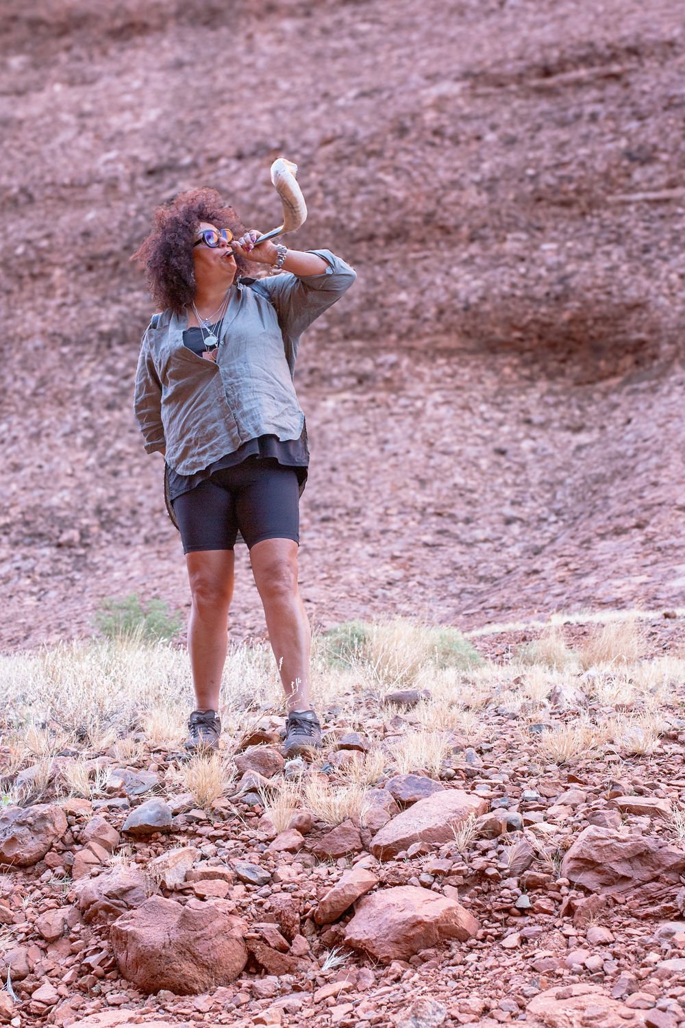 A person with curly hair and glasses standing on rocky terrain, holding a large bone-shaped object to their mouth as if blowing into it, with a reddish cliff in the background.