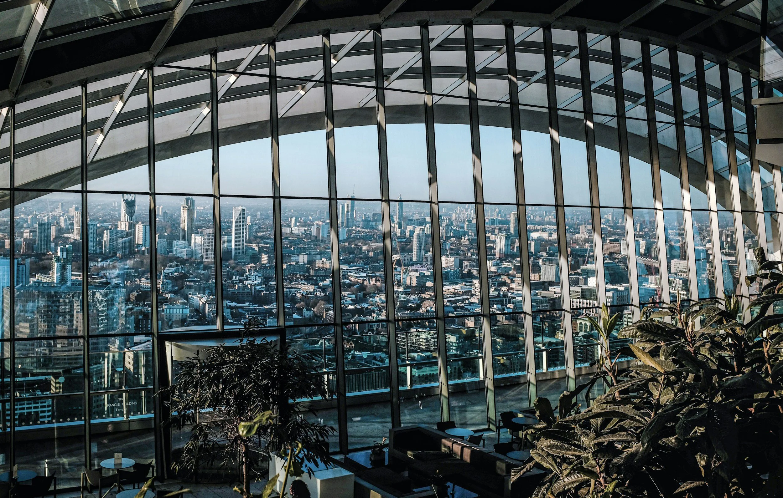 View of a city skyline through large glass windows in a high-rise building, with plants and seating visible in the foreground.