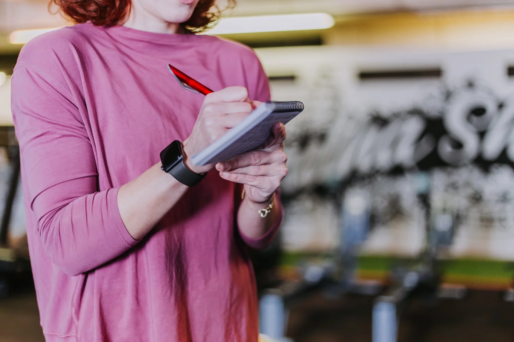 A woman wearing a pink long sleeve shirt writing in a notepad with a red pen inside a gym or sports facility.
