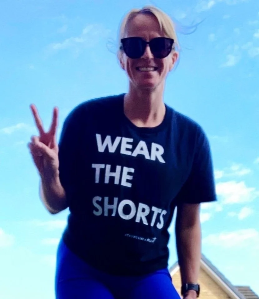 Woman smiling outdoors wearing sunglasses and a black T-shirt that says "Wear the Shorts". She is making a peace sign with her right hand and standing against a bright blue sky.