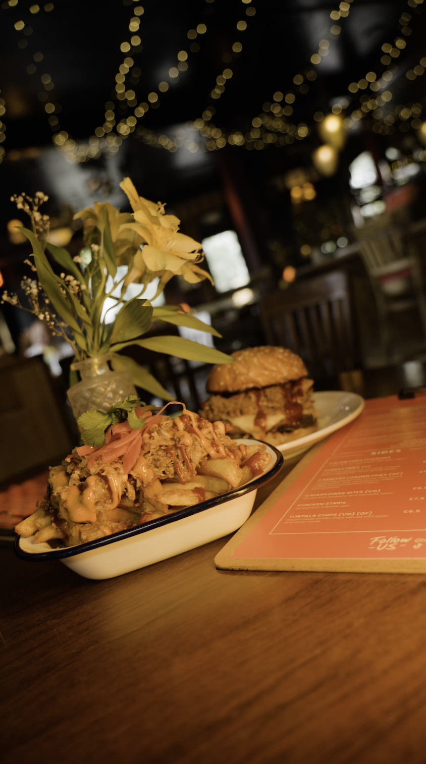 A plate of loaded fries with toppings near a picnic style burger on a white plate on a wooden table with a red menu, a vase with white flowers, and warm background lighting.