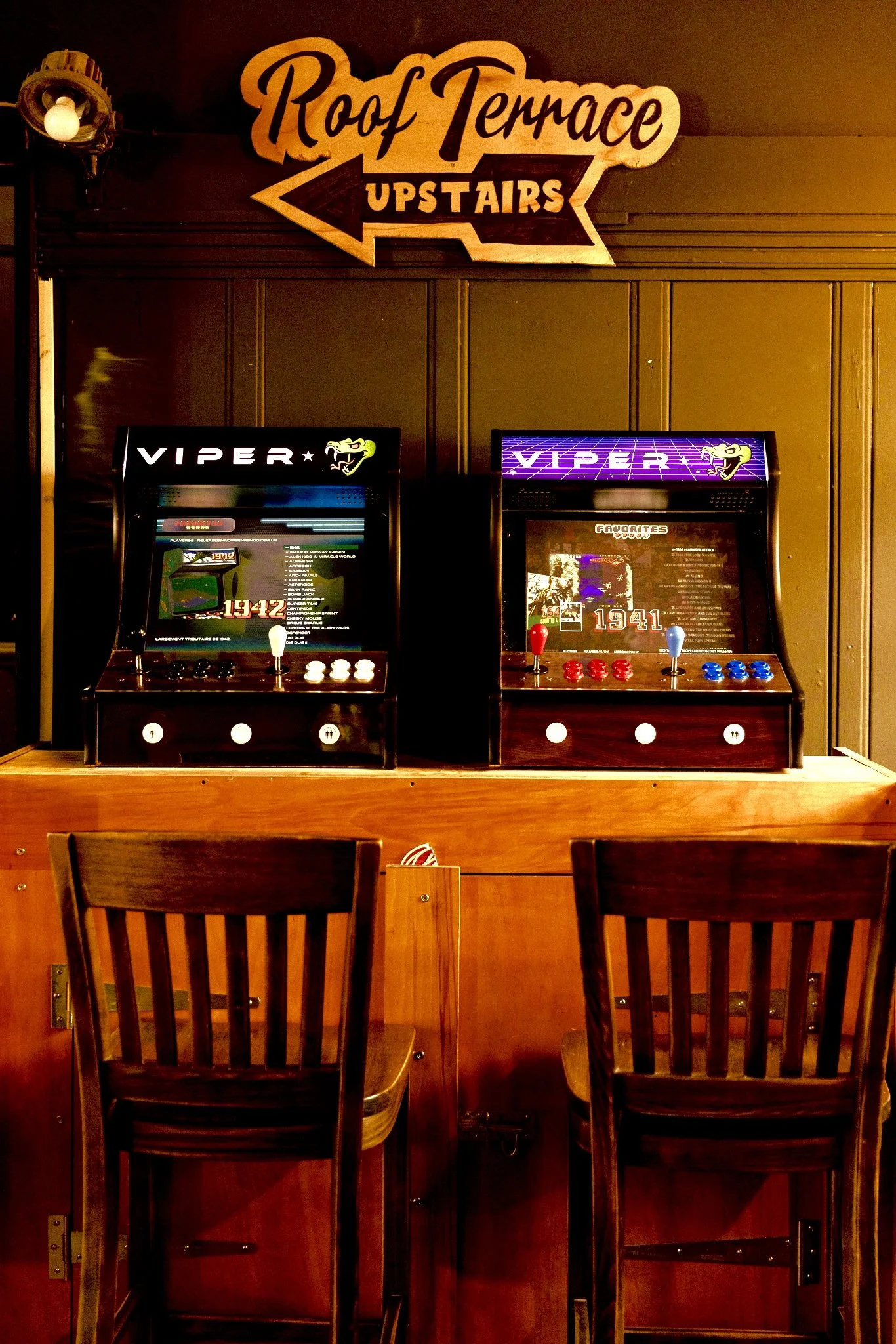 Indoor view of two vintage arcade game machines on a wooden counter, with wooden chairs in the foreground. A sign hanging above indicates the location is "Rooftop Terrace Upstairs."