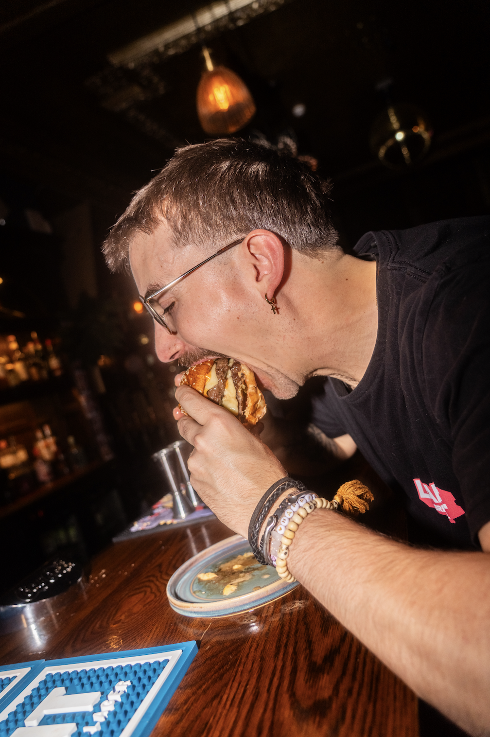 A man wearing glasses and bracelets taking a big bite out of a burger in a dimly lit bar or restaurant.