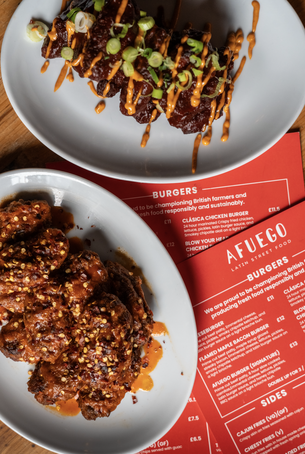 Two plates of food on a table, with a menu underneath. The top plate has glazed chicken wings garnished with chopped green onions and drizzled with orange sauce. The bottom plate contains fried chicken pieces topped with a spicy sauce and crushed pea