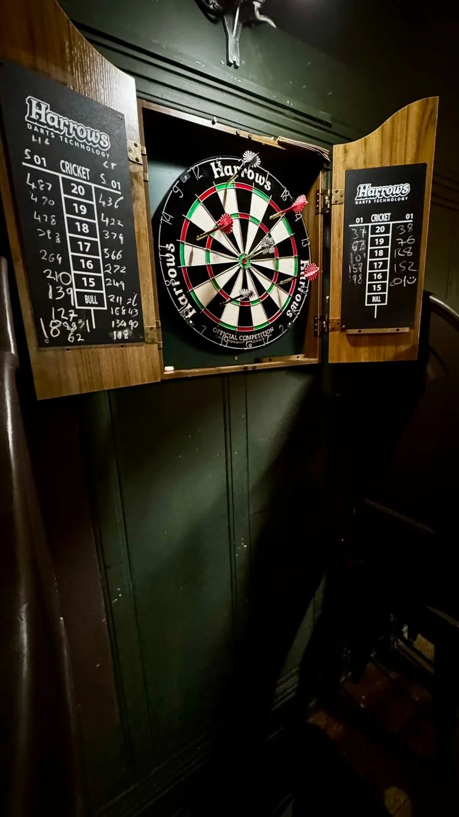 A dartboard in a pub with scoreboards on either side showing scores for cricket and standard darts. The dartboard has several darts attached, with some scoring on it.
