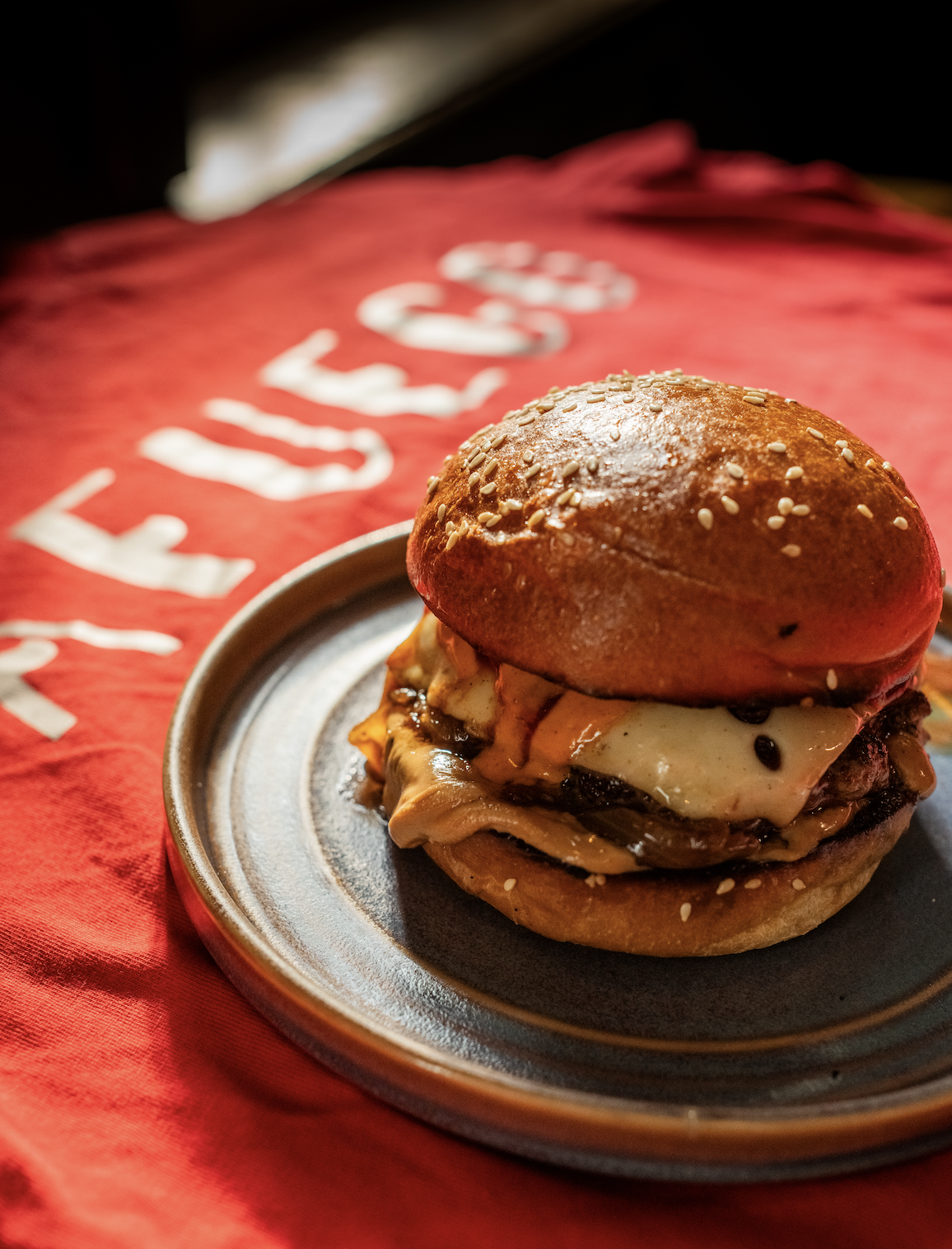 Close-up of a cheeseburger with a sesame seed bun, melted cheese, and grilled beef patty on a small dark plate, with a red cloth background.