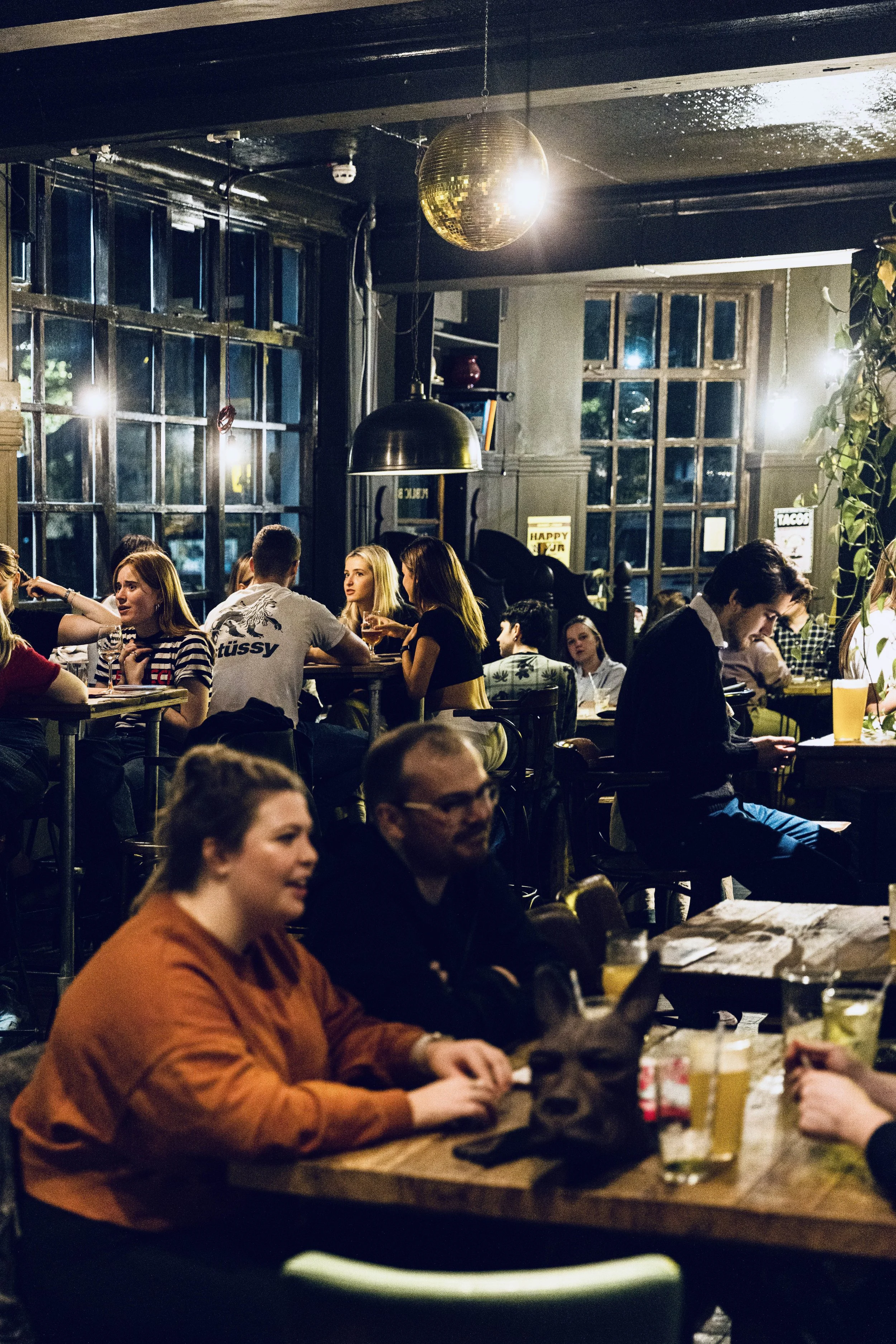 People dining and socializing inside a dimly lit restaurant with large windows, a disco ball hanging from the ceiling, and a mix of casual and semi-formal attire.
