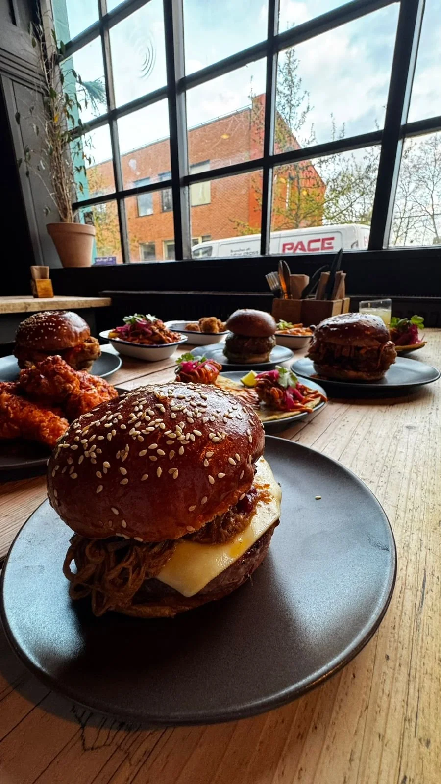 A variety of burgers and side dishes on a wooden table near a large window with a view of a brick building and trees outside.