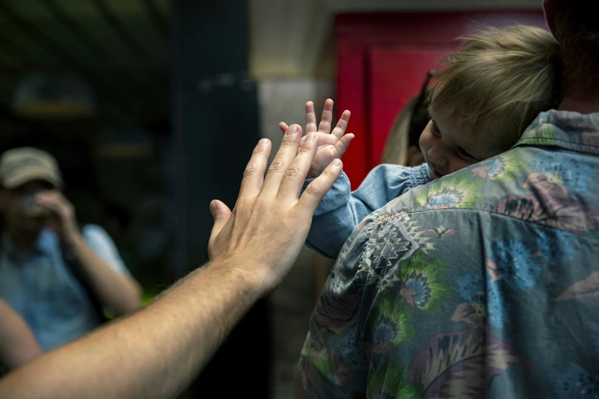Child with blonde hair receiving a high-five from an adult with a tattooed arm, wearing a colorful patterned shirt, in a dimly lit indoor setting.