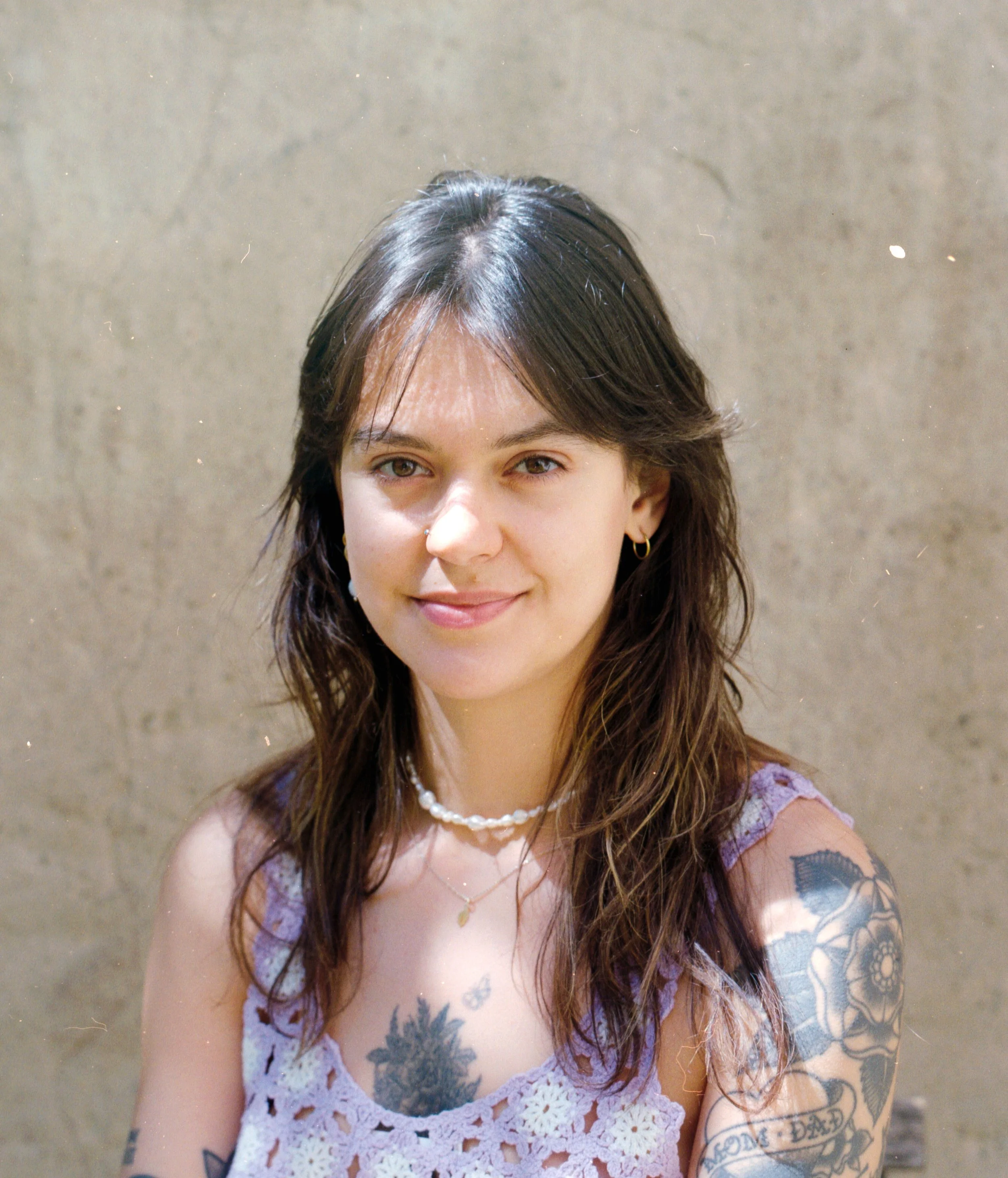 Close-up of a woman with brown hair and hazel eyes, smiling and wearing a lilac lace dress, gold hoop earrings, a pearl necklace, and tattoos on her arm, standing in front of a beige wall. Doula, psychologist, teacher, counselor, birth-worker.