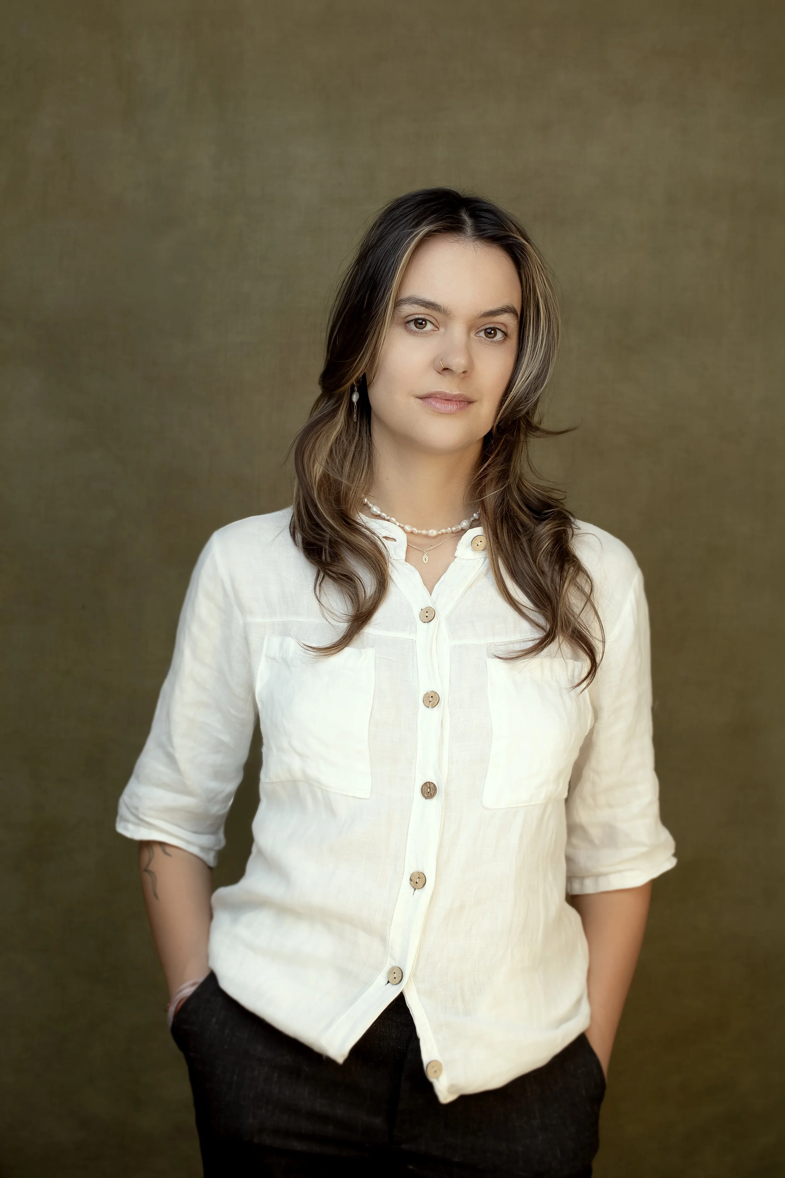 A woman with shoulder-length brown hair wearing a white button-up shirt, black pants, and jewelry standing against a brown background.