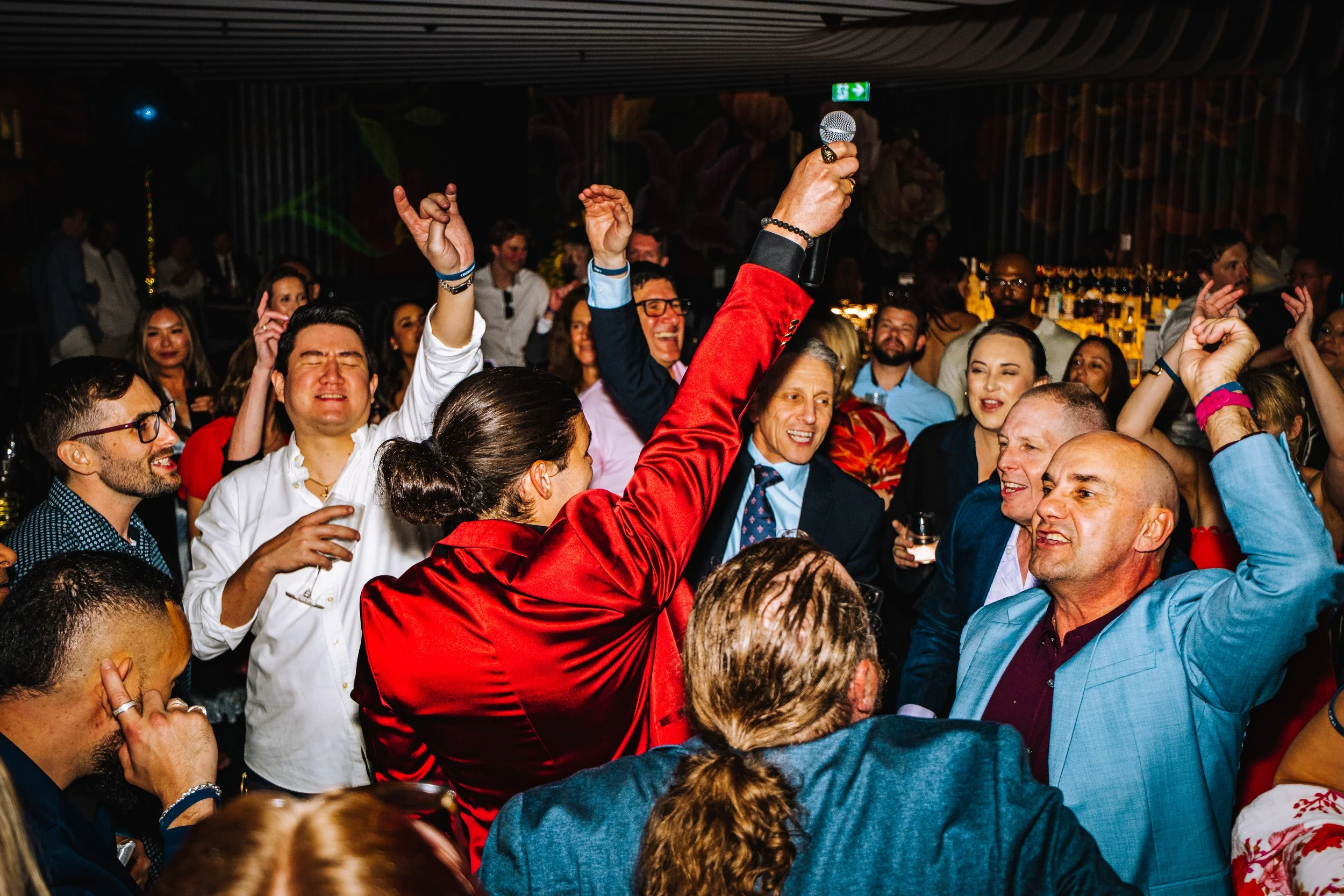 Crowd of people at a lively party, dancing and enjoying music, with a person in a red jacket holding a microphone in the center.