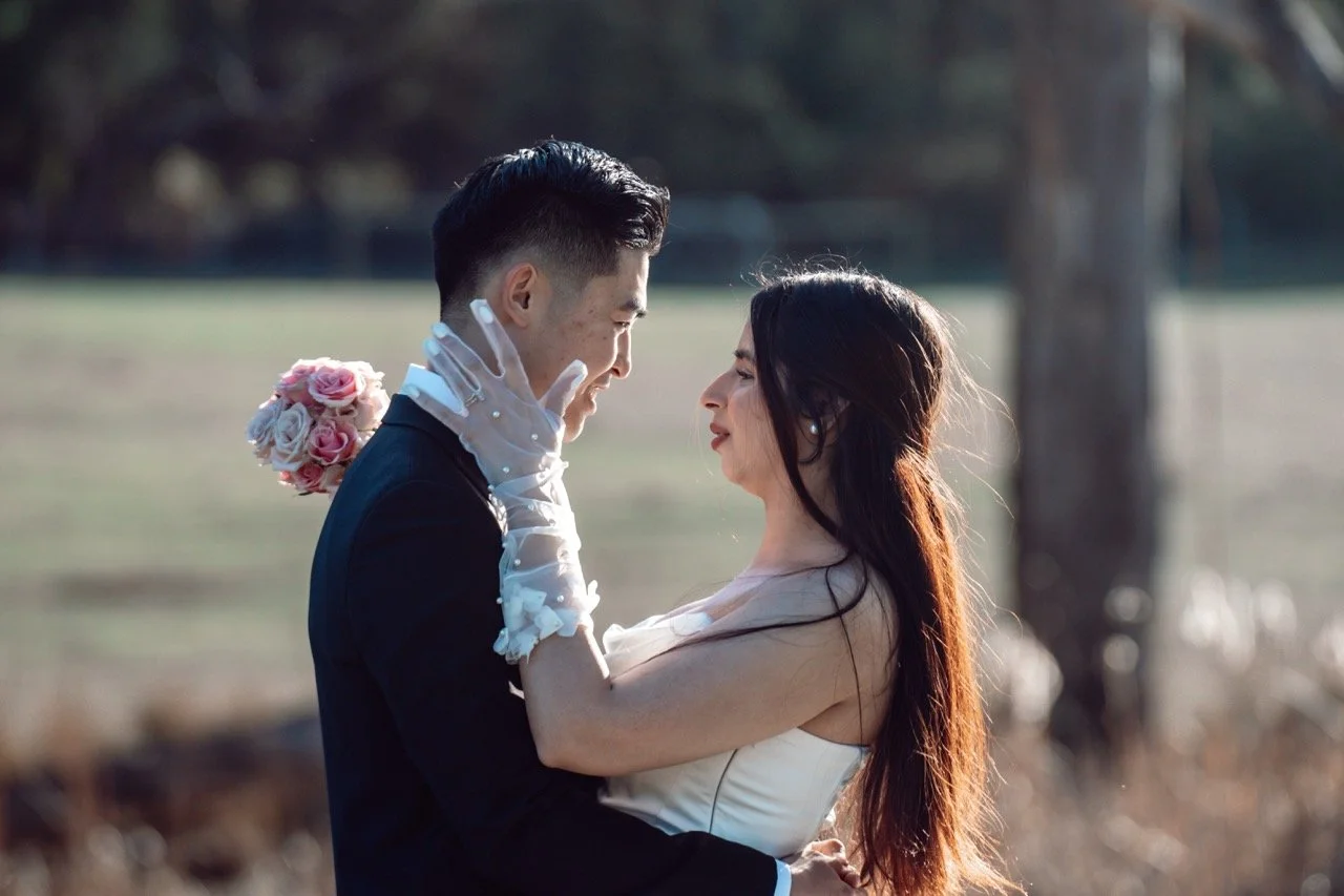 A couple in wedding attire smiling and holding each other outdoors, with trees in the background.