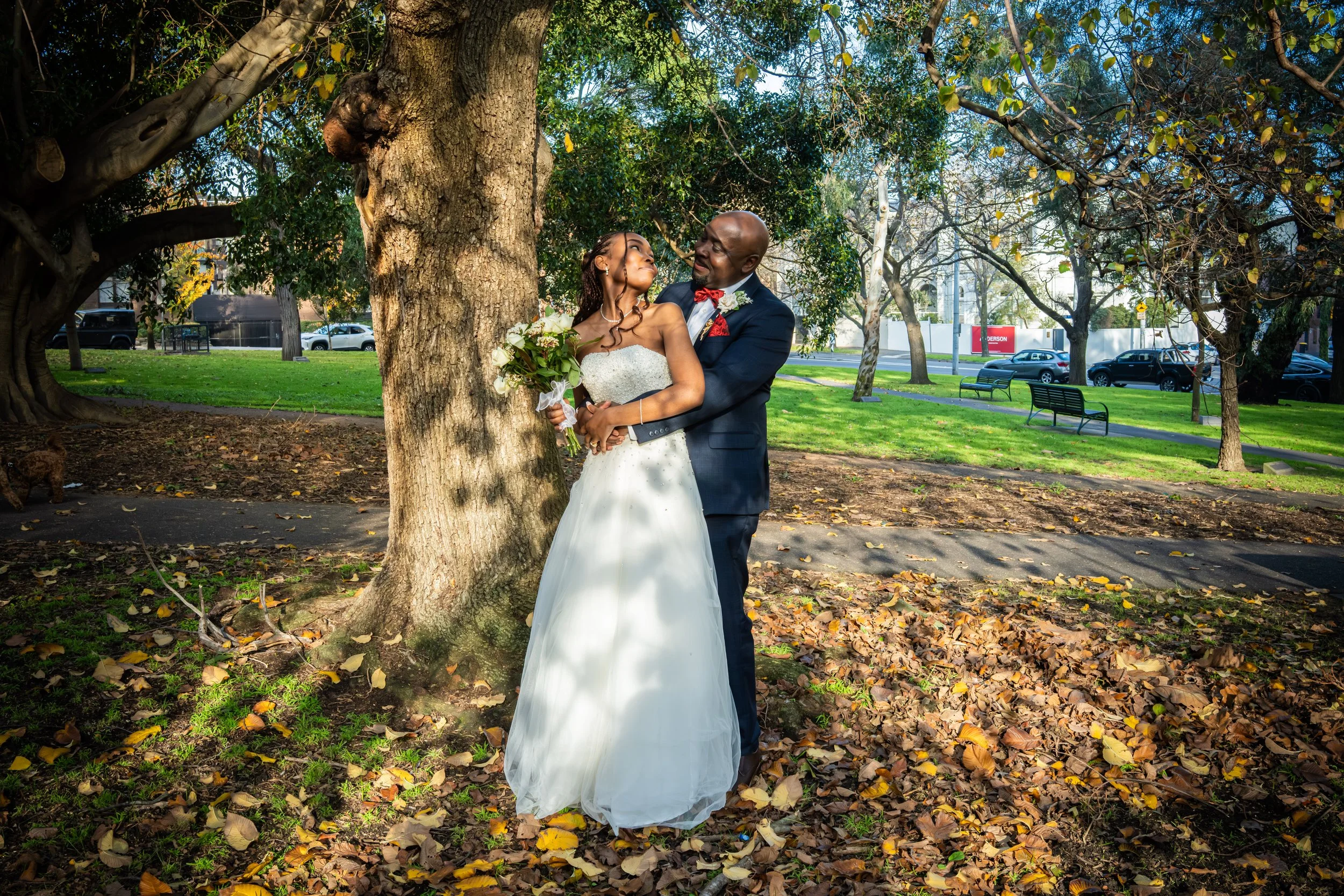 A bride and groom in wedding attire standing outdoors among trees and fallen leaves, smiling and looking at each other. The bride is holding a bouquet of flowers, and they are embracing near a large tree.