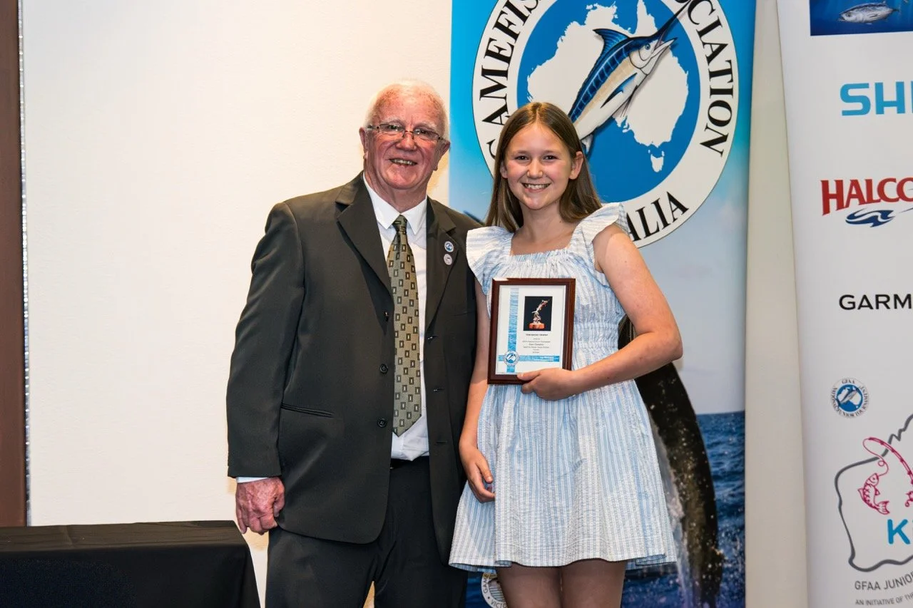 A young girl is smiling and holding a framed award while standing next to an older man in a suit. They are at an event with a large banner featuring a fish in the background.