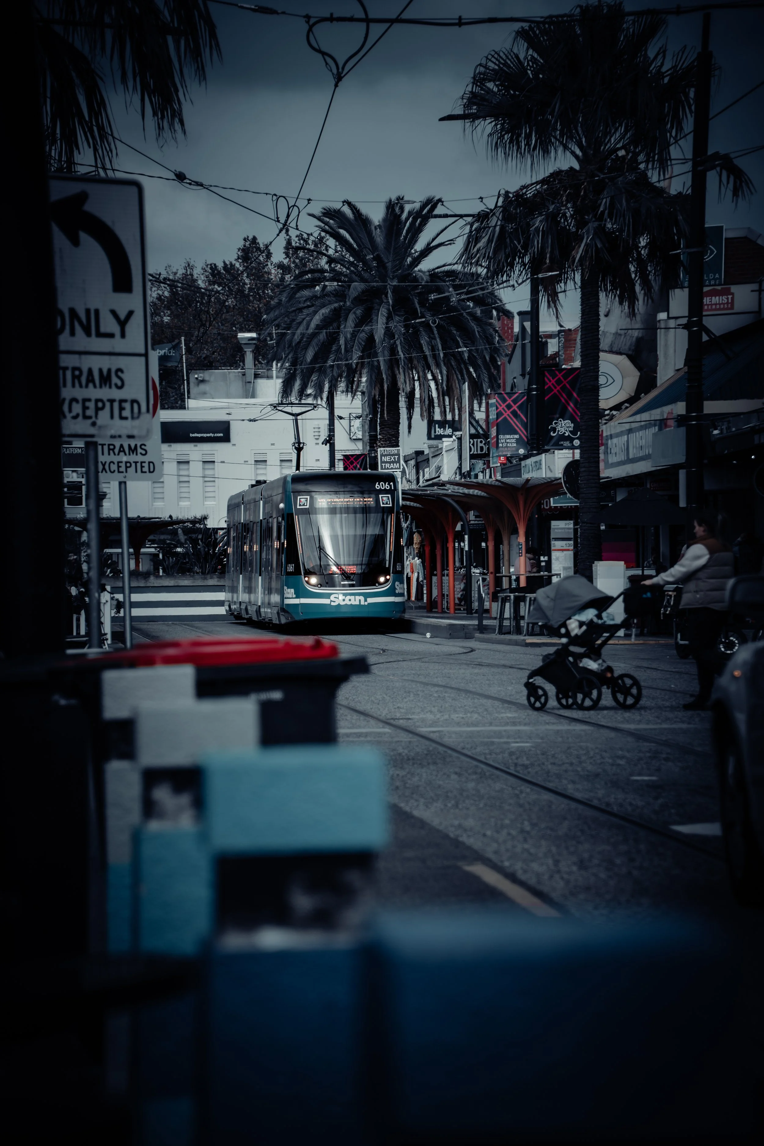 An urban street scene with a modern tram approaching a stop, palm trees, and storefronts. A person is pushing a stroller on the sidewalk, and there are signs and overhead wires visible.