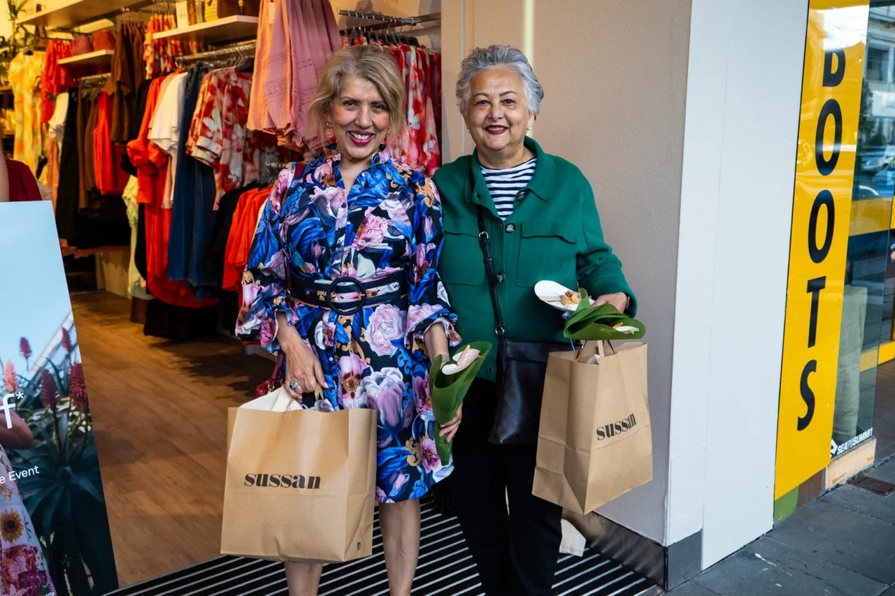 Two smiling women standing outside a clothing store, holding shopping bags and flowers, with colorful clothes displayed inside the store.