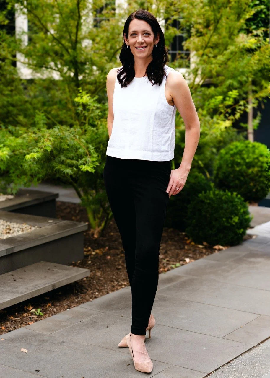 A woman standing outdoors on a sidewalk, smiling, wearing a white sleeveless top, black pants, and beige high heels, with green trees and bushes in the background.