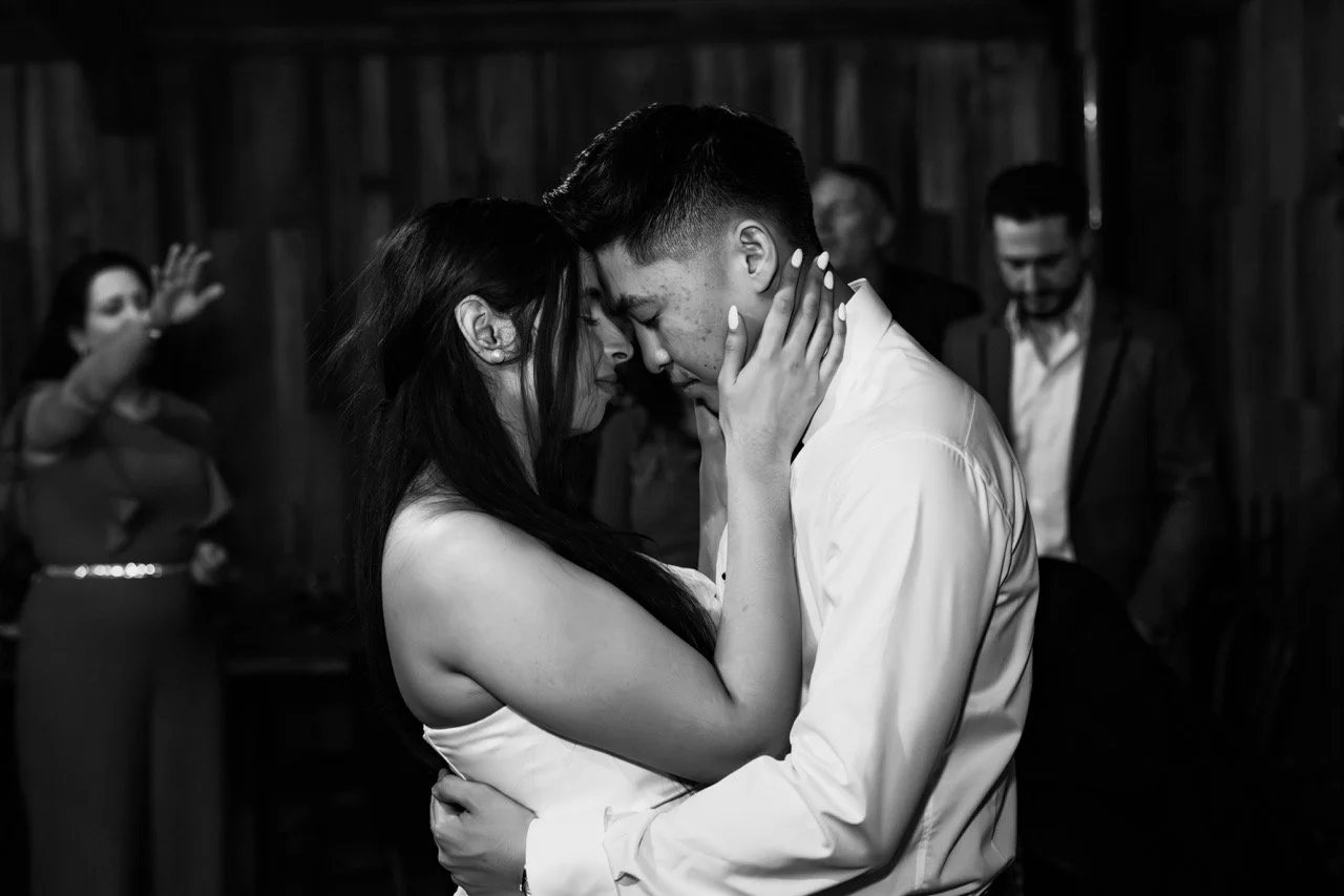 A couple dances closely with foreheads touching in a black and white photo, while three people watch in the background.