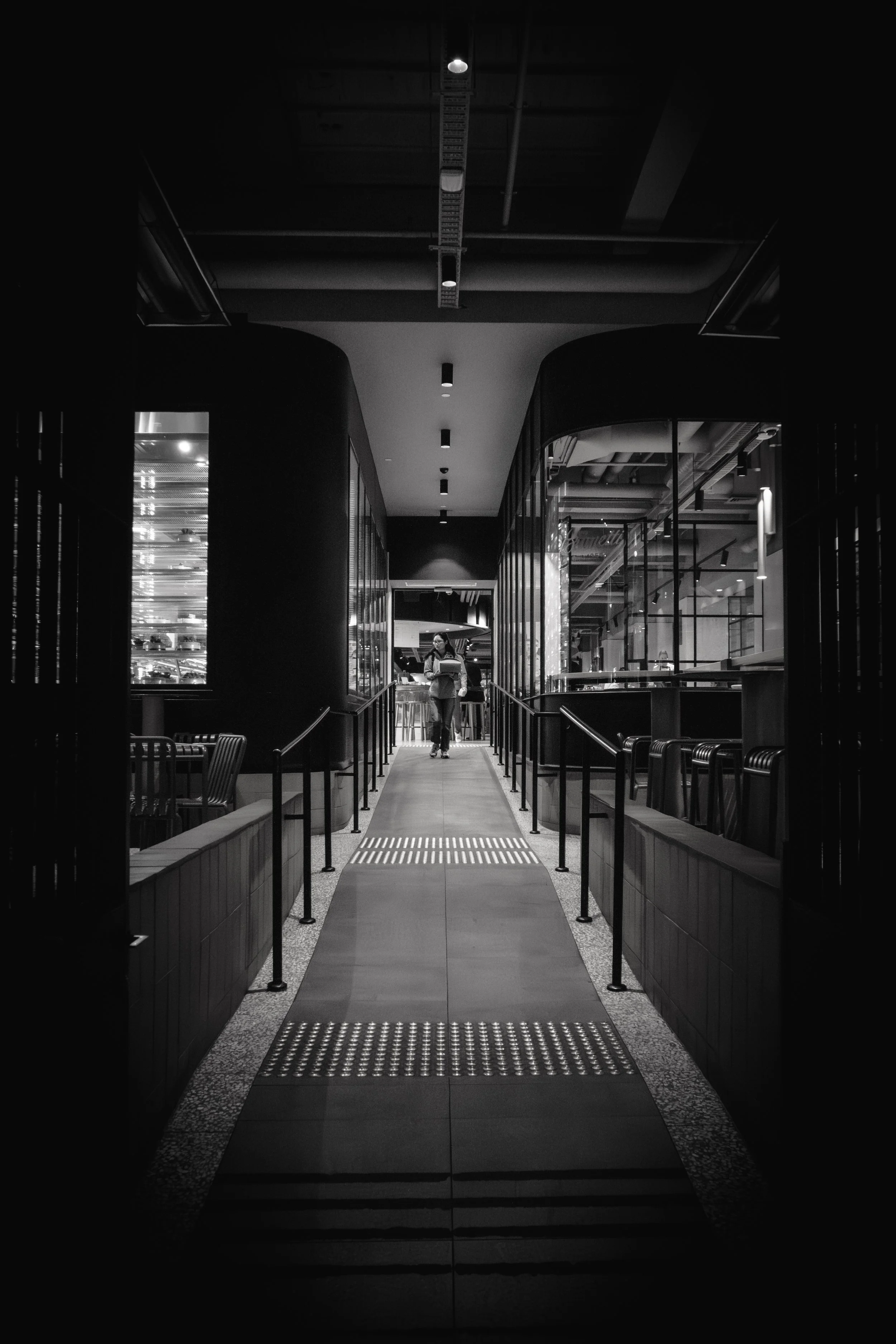 Black and white photo of a modern indoor corridor with metal railings, wooden benches, and glass walls, leading to a person walking in the distance.
