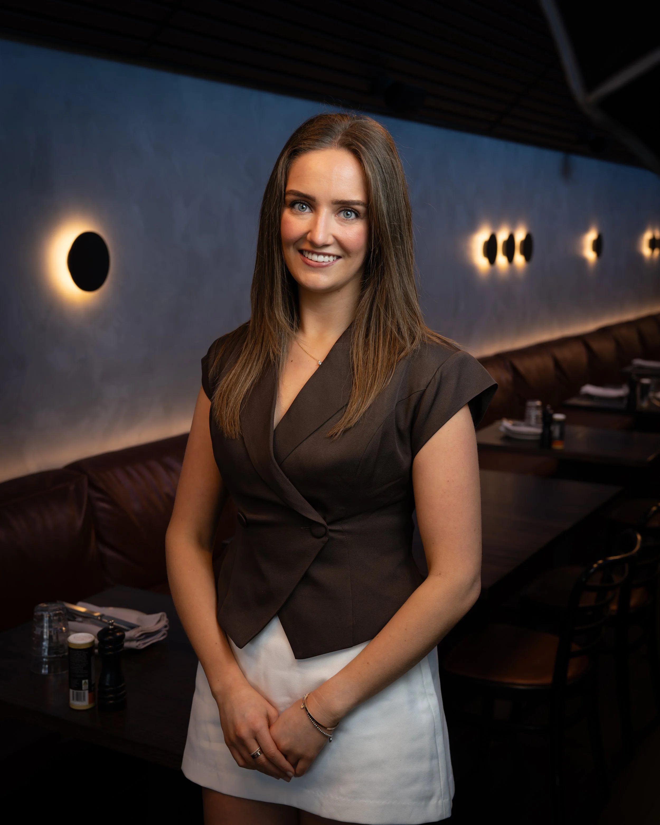 A young woman with long brown hair, blue eyes, and wearing a dark brown blazer over a white dress, standing in a restaurant or cafe with dim lighting and round wall sconces.