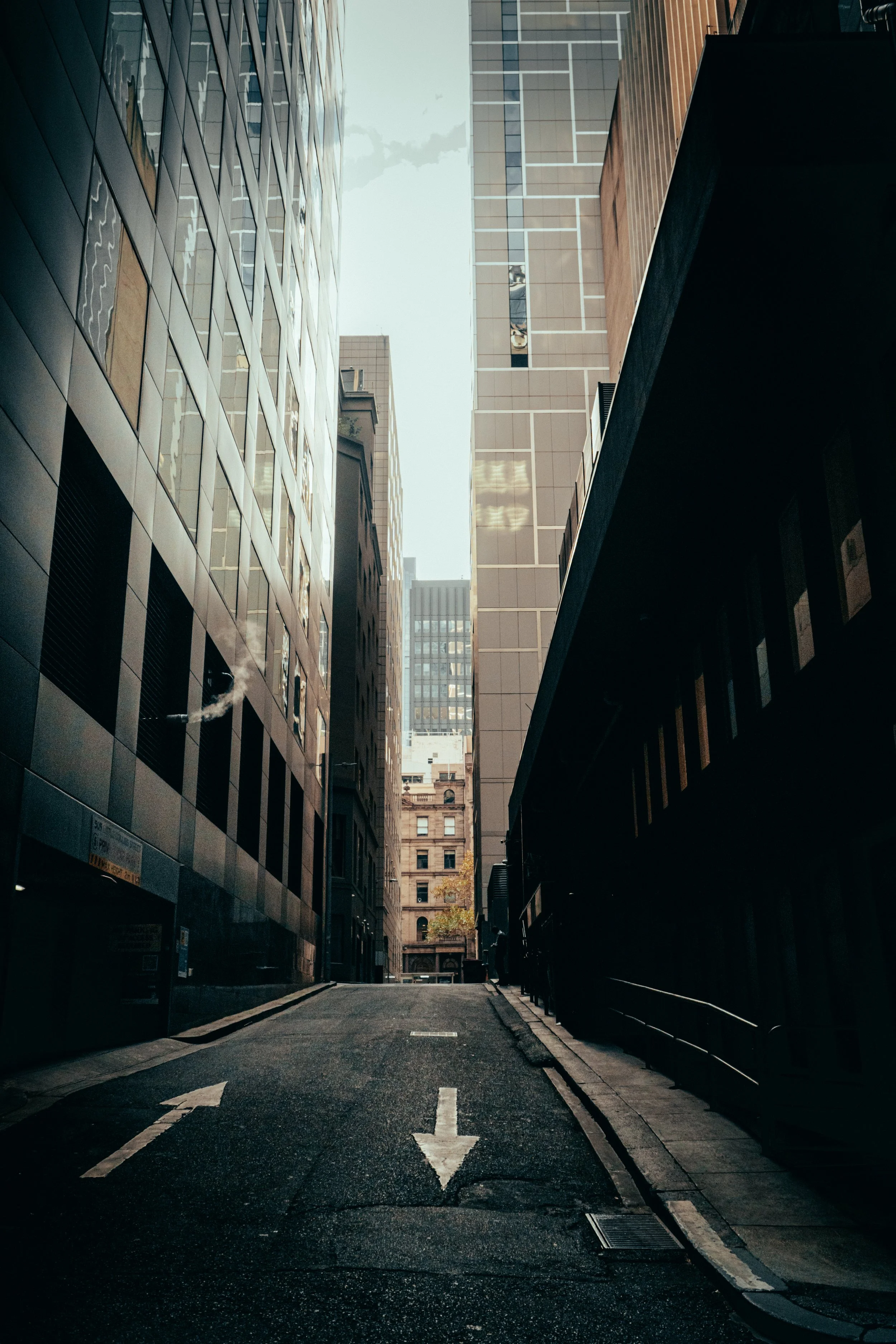 A narrow city street downhill between tall office buildings with glass and concrete facades, under a cloudy sky.
