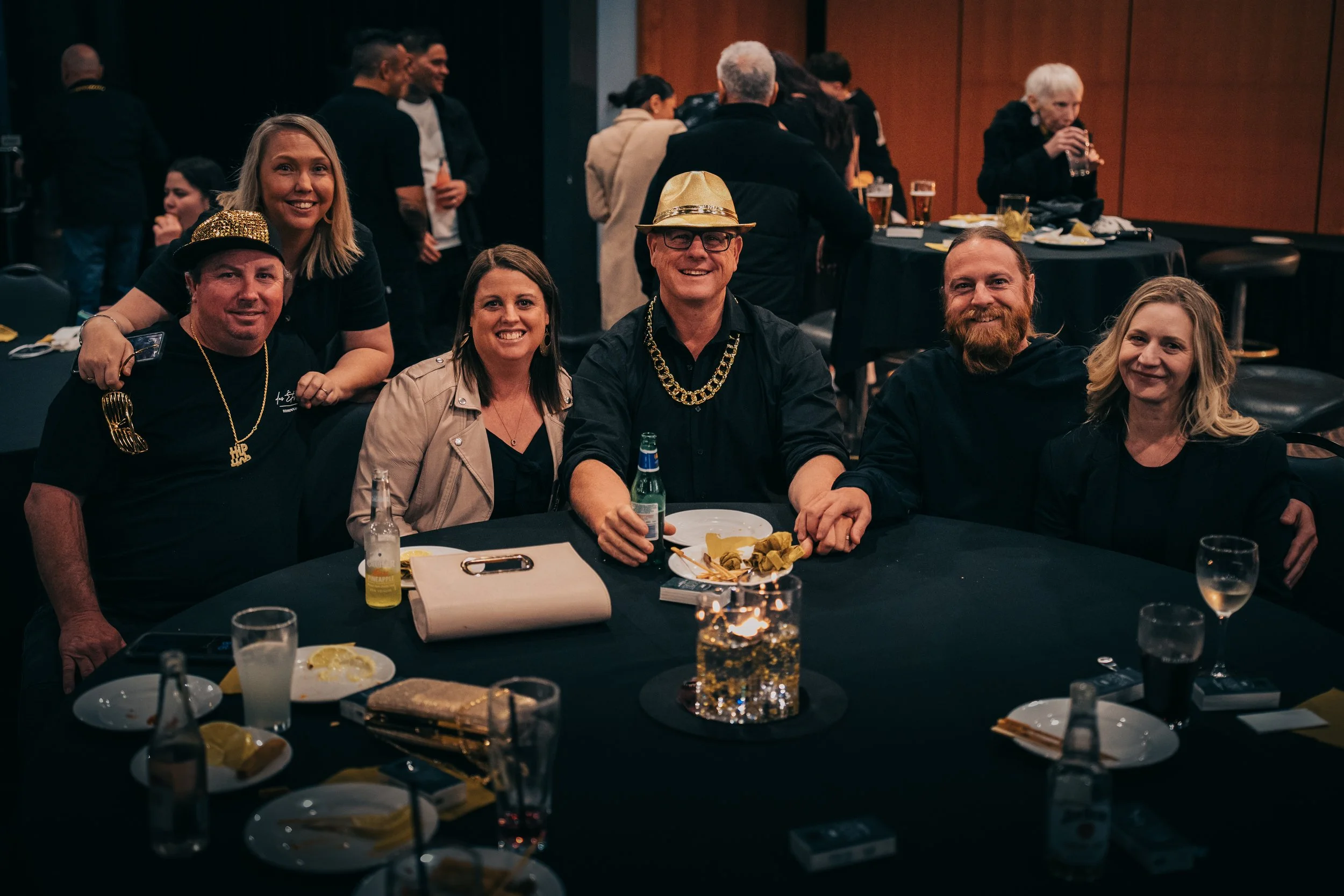 Group of six people at a party sitting around a table, with some wearing festive jewelry and hats, and others holding drinks, smiling for the camera.