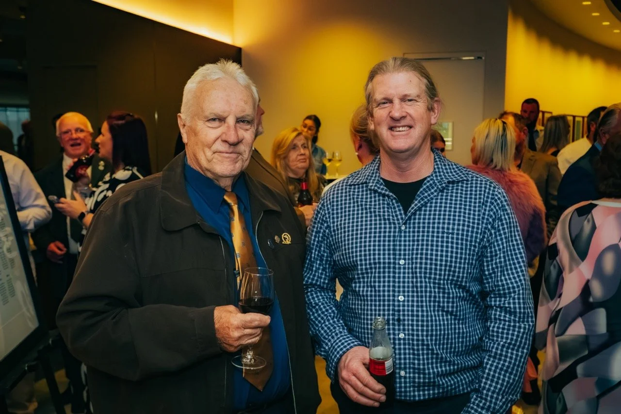 Two men at a social event, one older with white hair holding a glass of red wine, the other younger with blond hair holding a bottle of soda, smiling at the camera, with a crowd of people in the background.