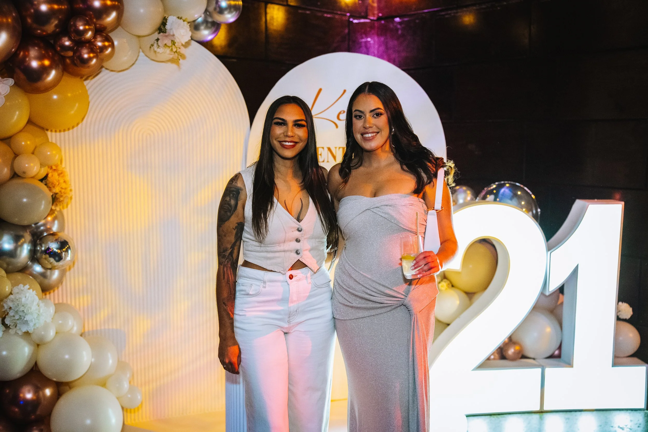 Two women standing together at a celebration, one holding a drink, in front of a decorative backdrop with balloons and a large illuminated '2021' sign.