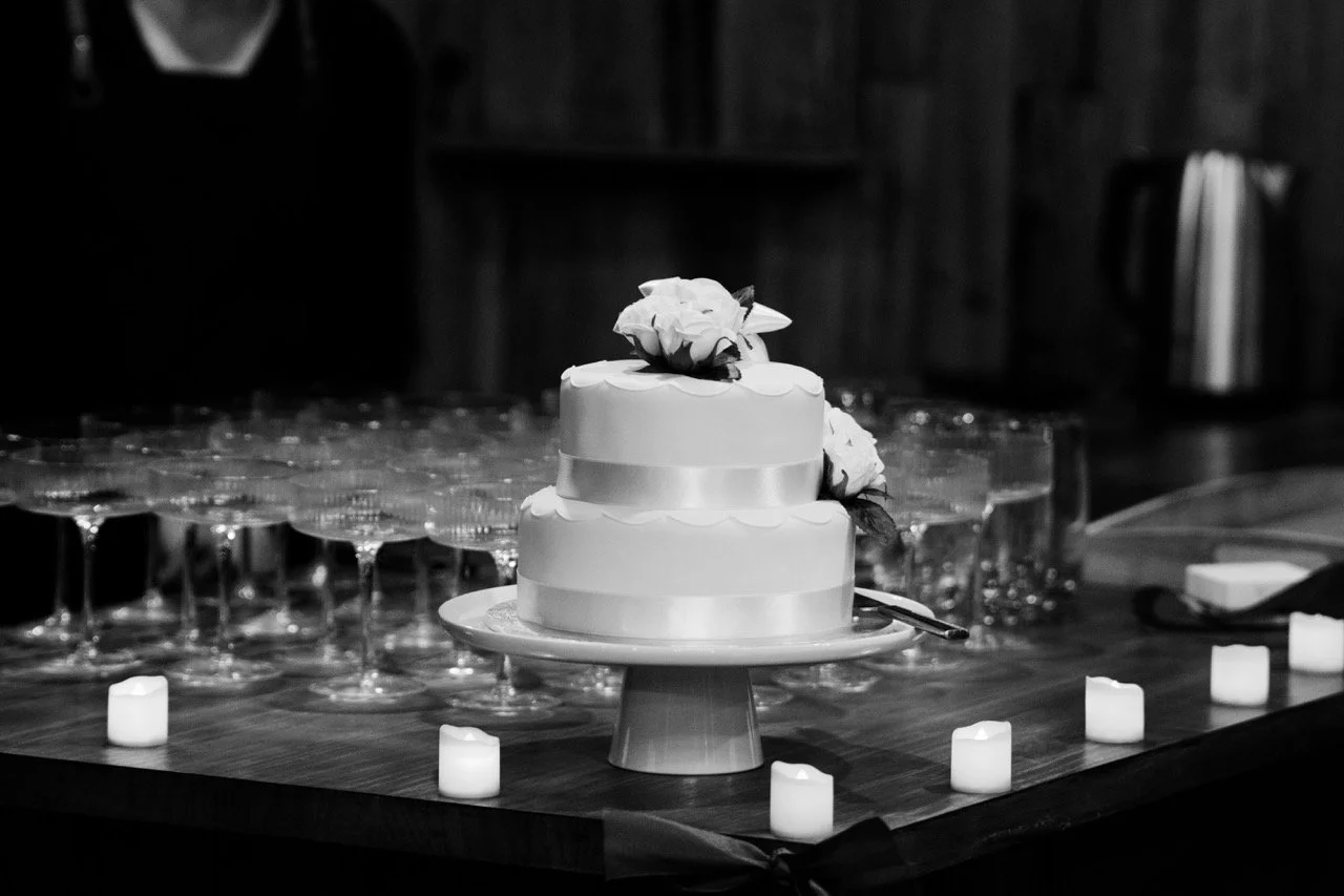 A two-tiered wedding cake with floral decorations on top, placed on a stand on a table. The table has several empty wine glasses and small LED candles around the cake. The background is dark with a wooden wall.