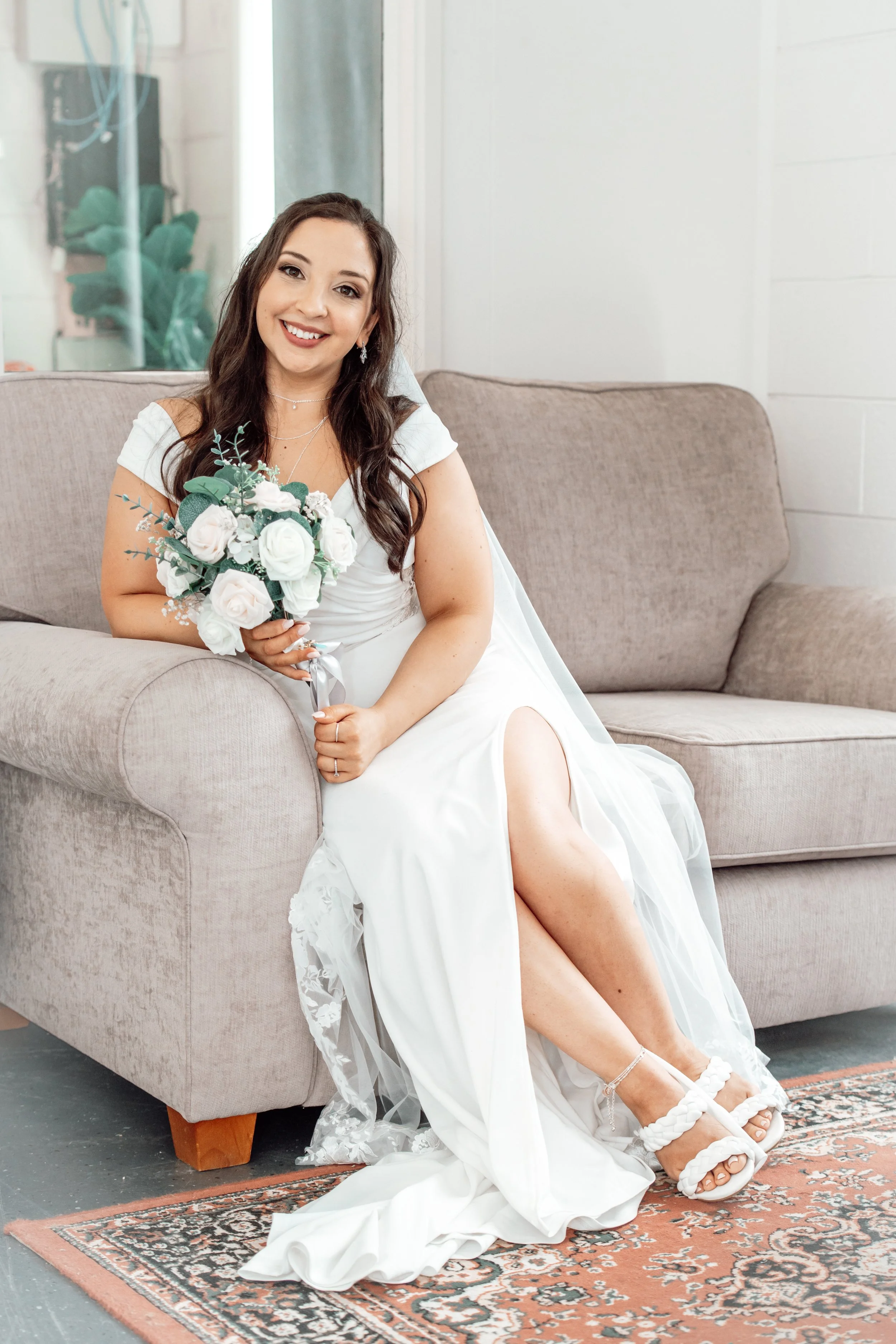 A woman in a white wedding dress sitting on a gray couch, holding a bouquet of white roses, smiling.