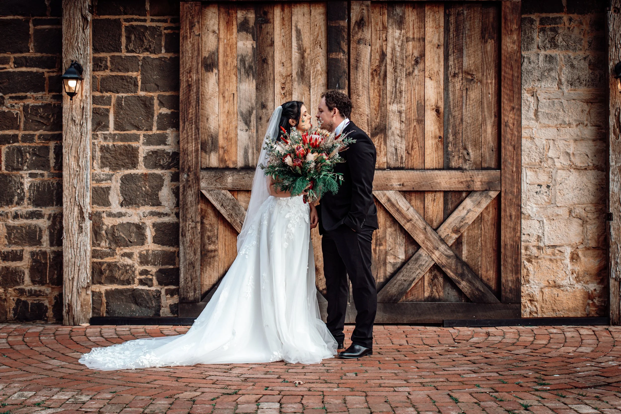 Bride and groom sharing a kiss in front of a rustic wooden barn door, with the bride holding a large bouquet of red, white, and green flowers, dressed in a white gown with a long train and veil, and the groom in a black suit and tie.