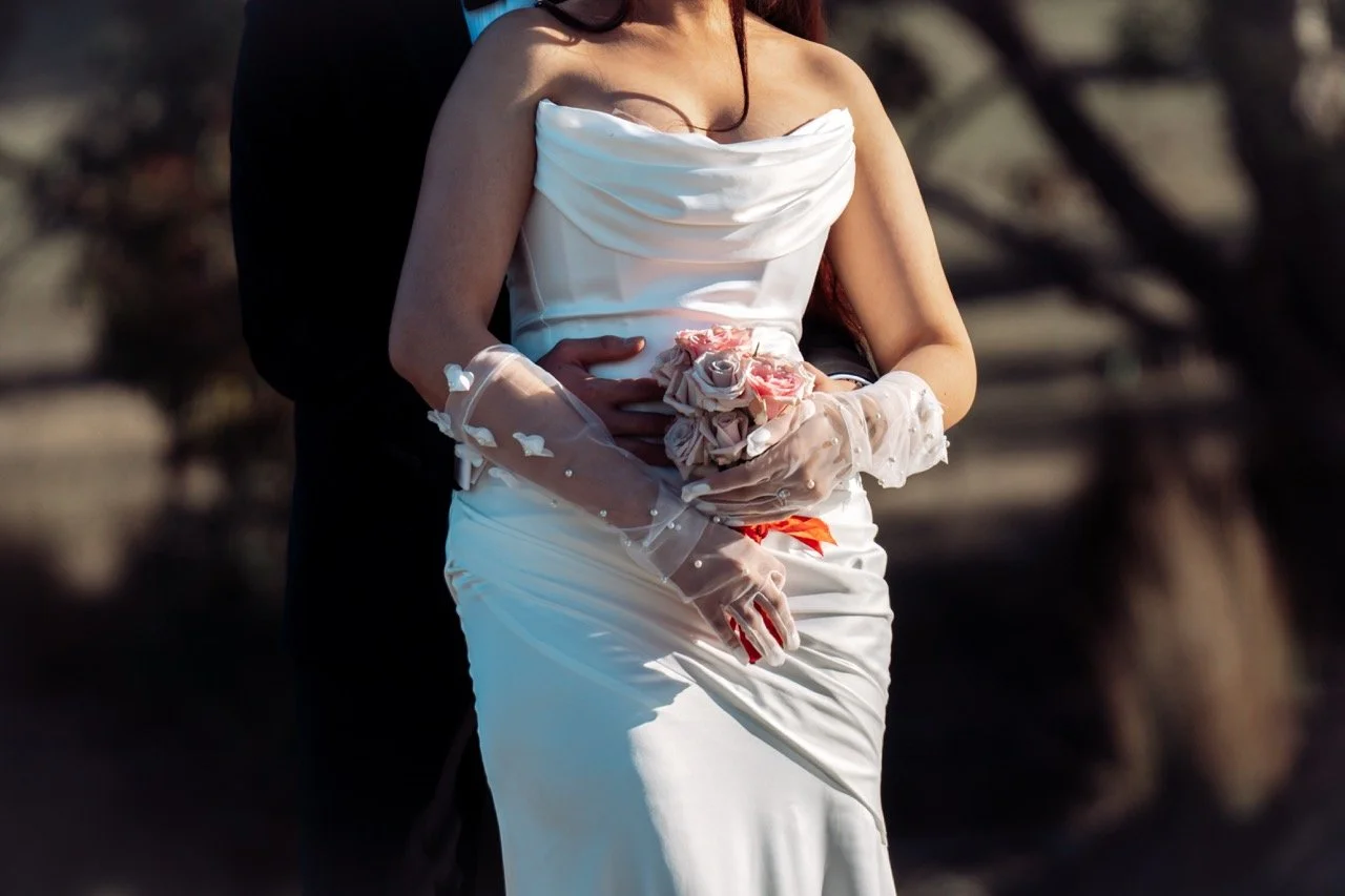 A bride in a white strapless wedding dress holding a bouquet of pink and white roses, standing close to a groom whose arms are around her waist, outdoors with blurred background.