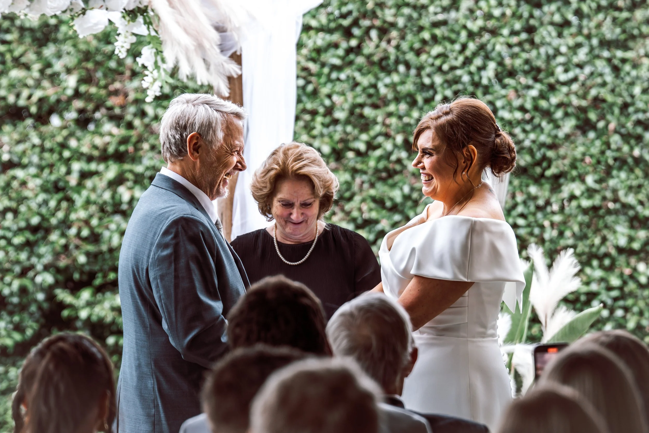 A bride and groom exchanging wedding vows with an officiant during a wedding ceremony outdoors, with friends and family witnessing, against a lush green background.
