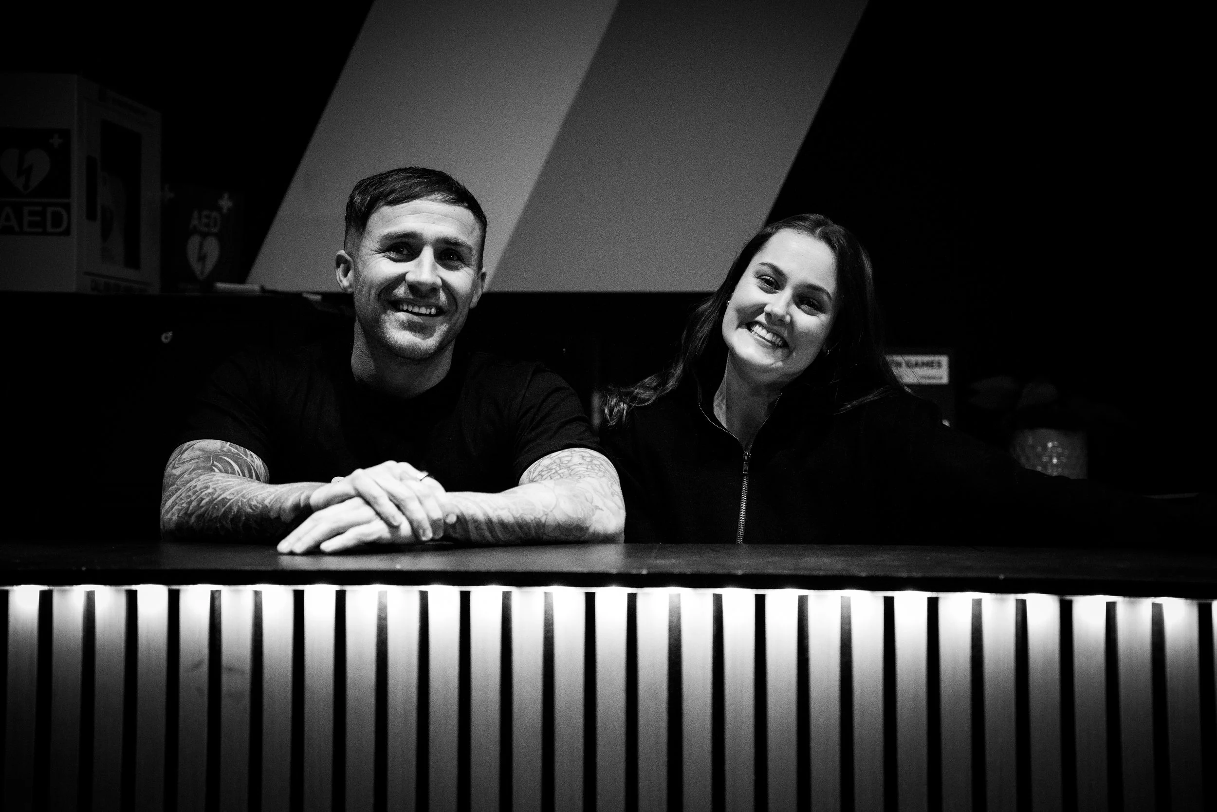 A man and woman smiling while sitting behind a counter in a dimly lit environment, with AED boxes visible in the background.