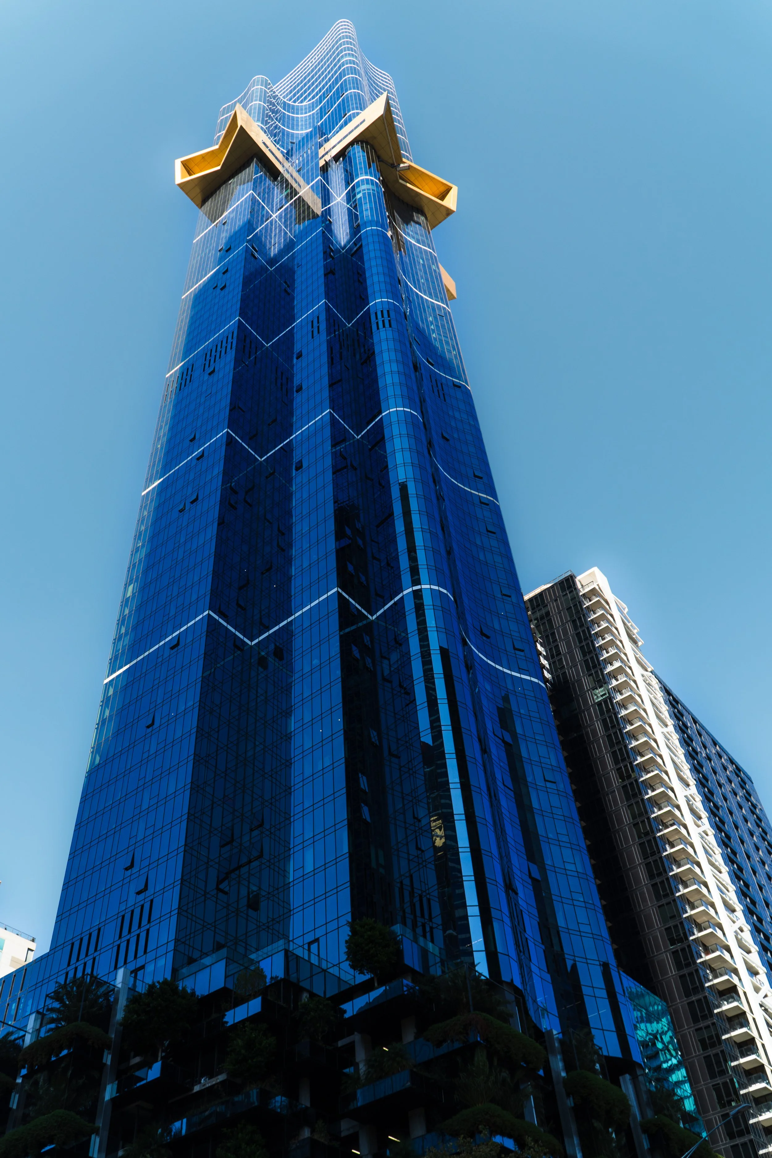 A tall modern skyscraper with a blue glass exterior, distinctive white lines, and yellow architectural features at the top, set against a clear blue sky.