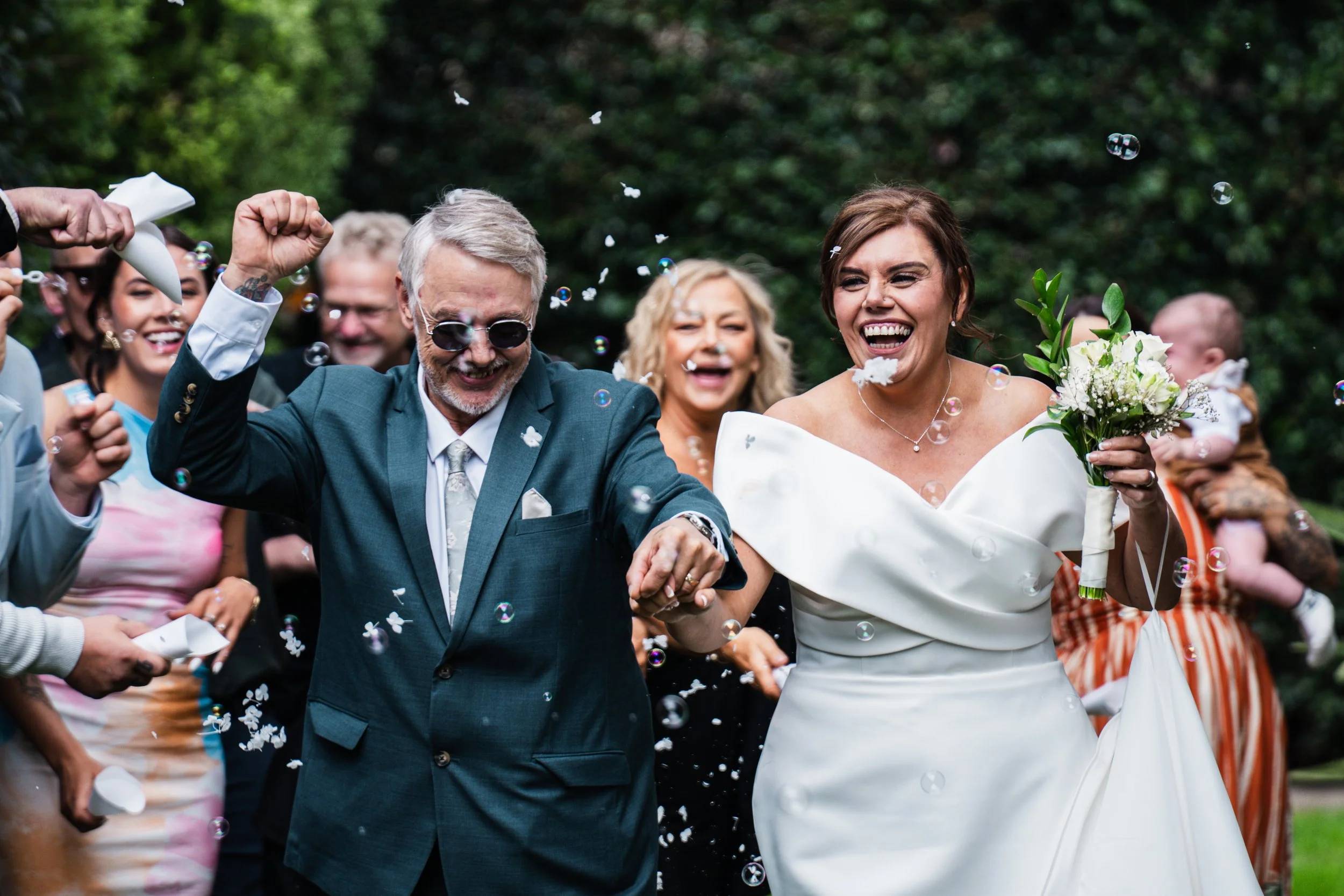 A bride and a groom, both smiling and holding hands, walking through a crowd of happy guests at a wedding celebration, with bubbles in the air and a lush green background.
