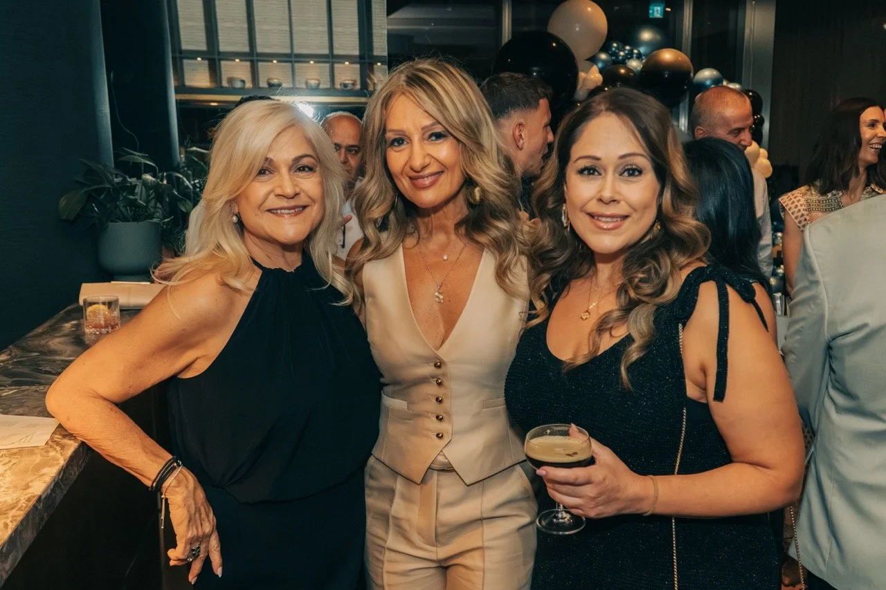 Three women at a celebration, smiling and dressed in elegant outfits, with balloons and other guests in the background.