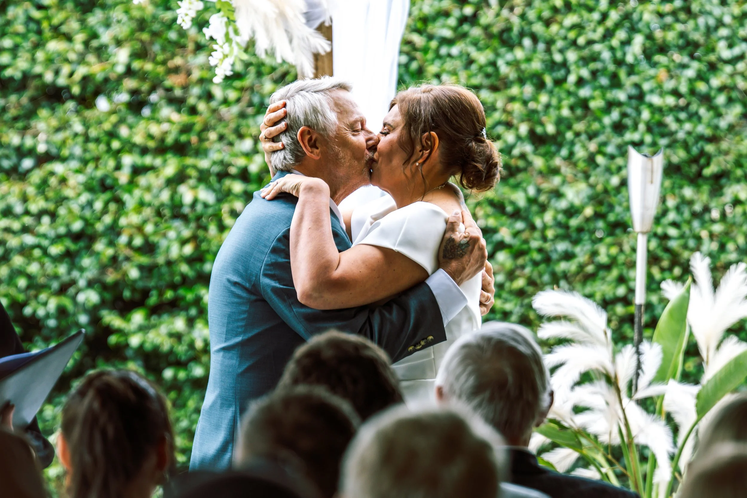 An elderly man and a woman share a romantic kiss during a wedding ceremony outdoors, surrounded by green foliage and white flowers.
