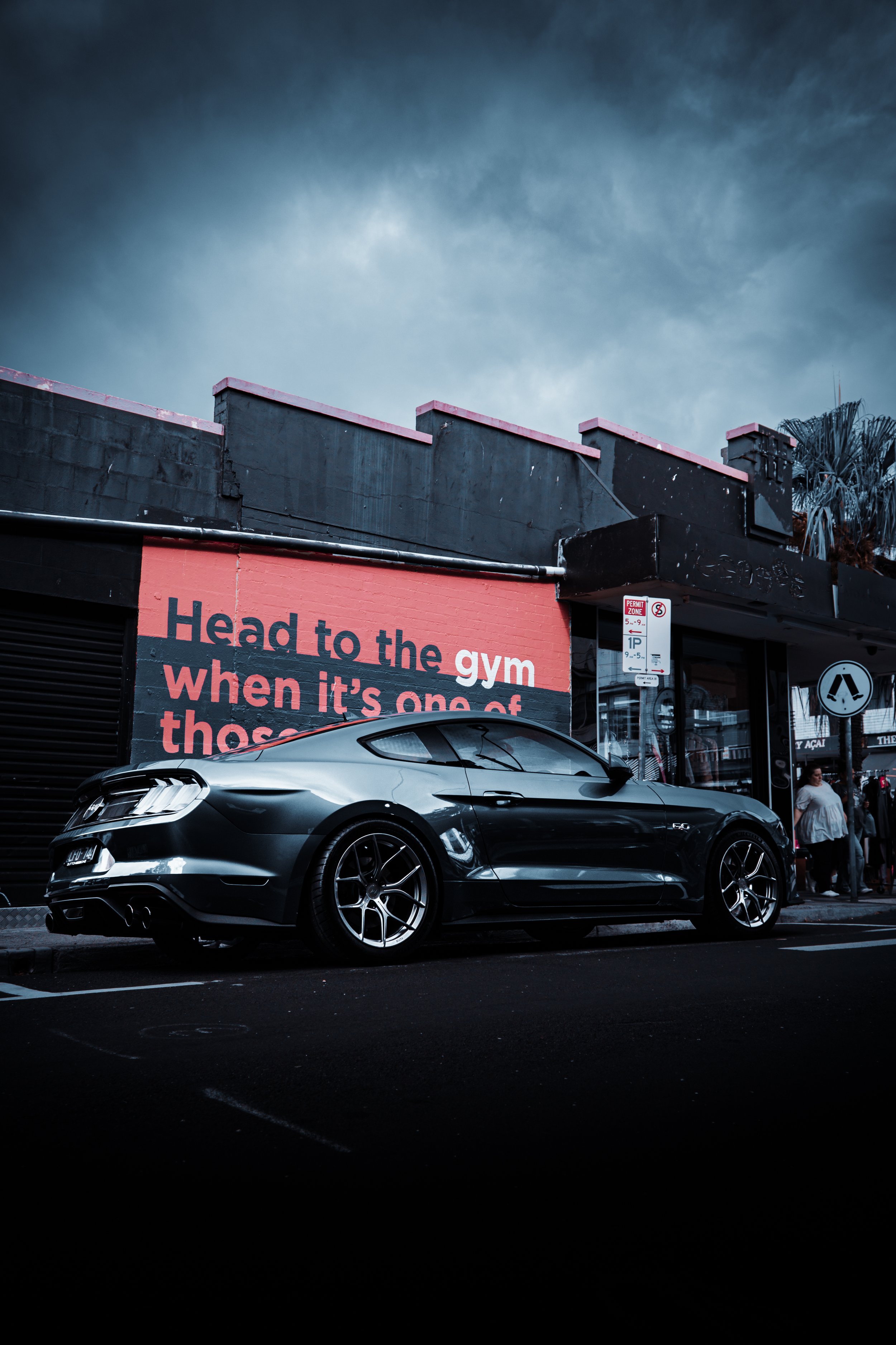 Black sports car parked on a city street at dusk, with a building with a mural that says 'Head to the gym when it's one of those' in pink and white. The sky is cloudy, and there are a few pedestrians nearby.